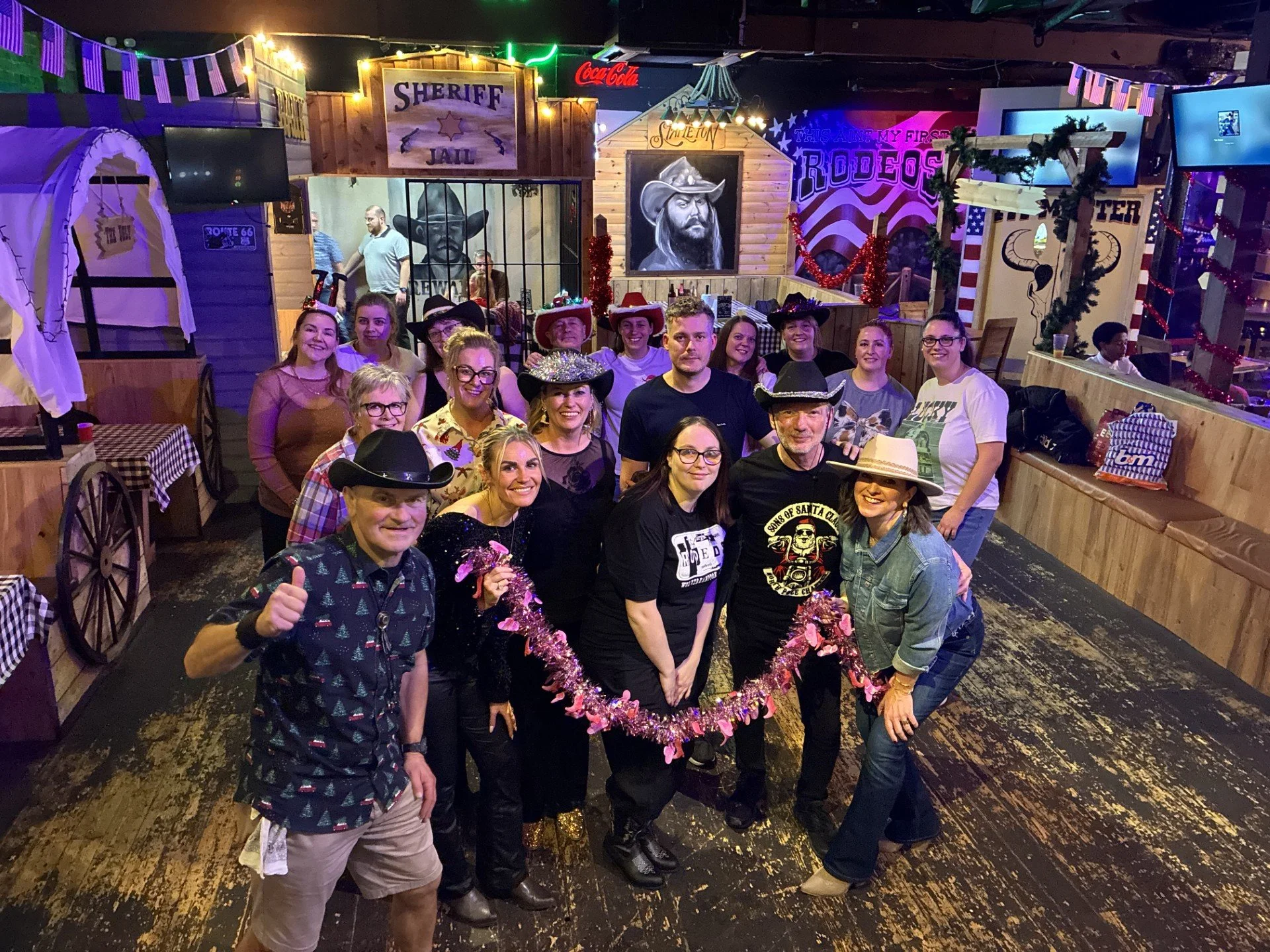 A group of people dressed in cowboy and cowgirl costumes poses in front of a western-themed backdrop with signs reading 'Sheriff', 'Jail', and 'Rodeo'. They are smiling and holding a pink garland, with some wearing hats. The background has decorations like string lights, banners, paintings of cowboys, and themed props.