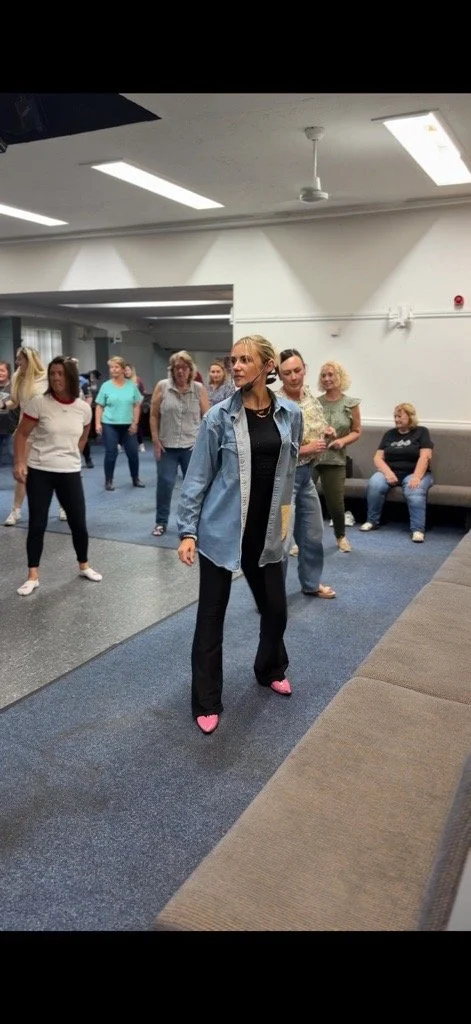 A group of women in a room, with one woman in the foreground wearing pink slippers, standing on a blue carpet and looking to the side.