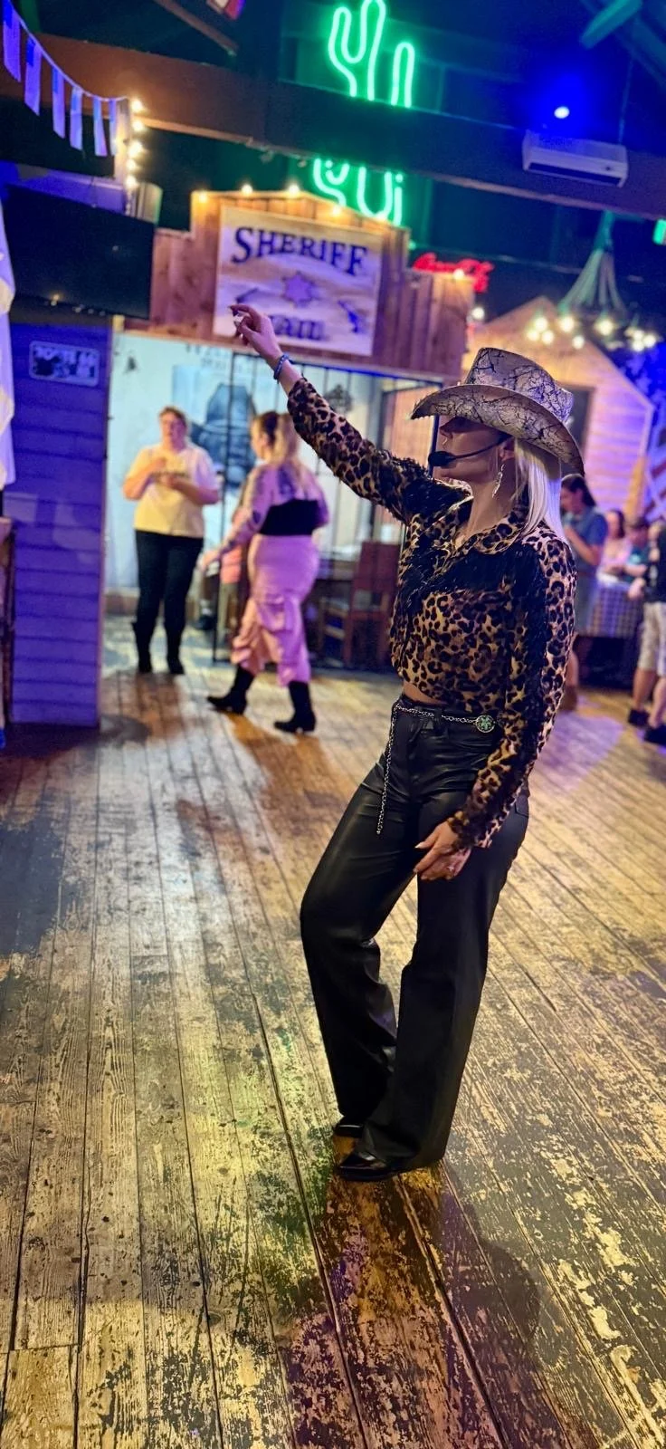 Woman wearing a leopard print top, black leather pants, and a cowboy hat posing and pointing in a Western-themed bar or restaurant with a neon cactus sign and sheriff sign in the background.