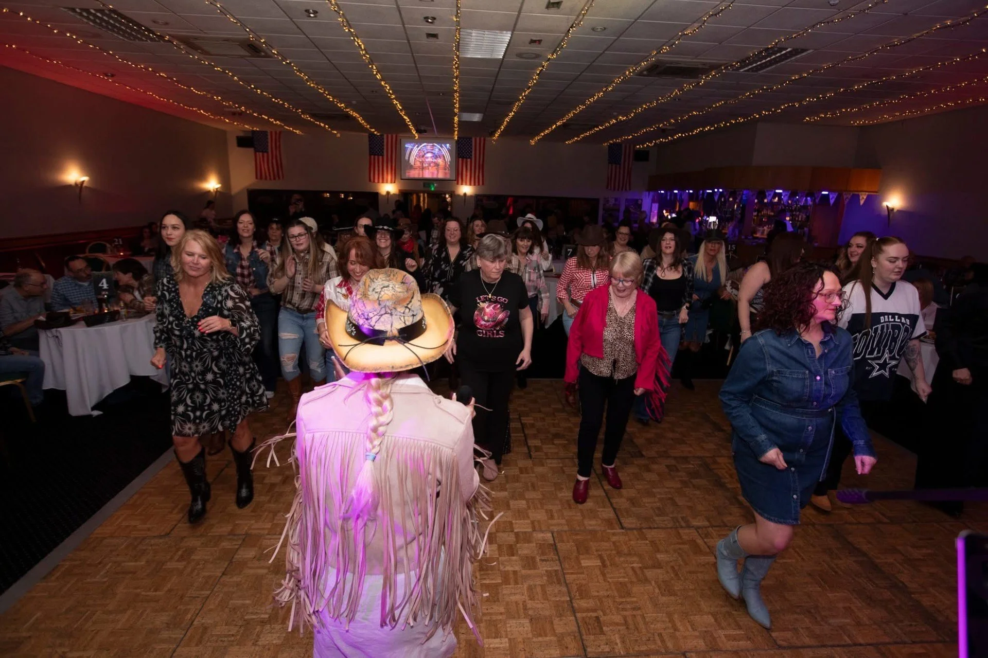 Women line dance at a country-themed event in a decorated hall with American flags and string lights, with a person in the foreground wearing a cowboy hat.