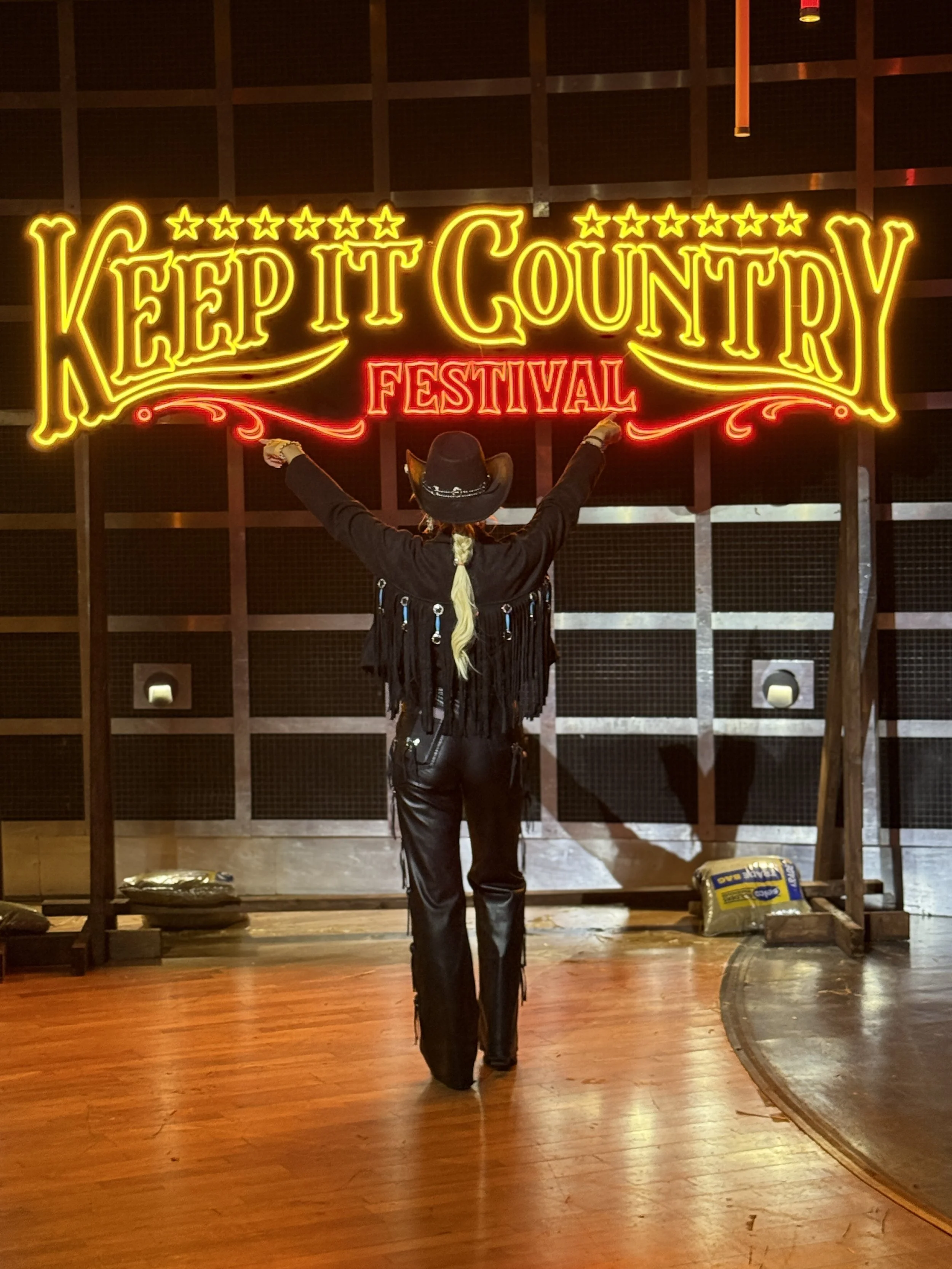 Person dressed as a cowboy standing in front of a neon sign that says 'Keep It Country Festival' with arms raised.