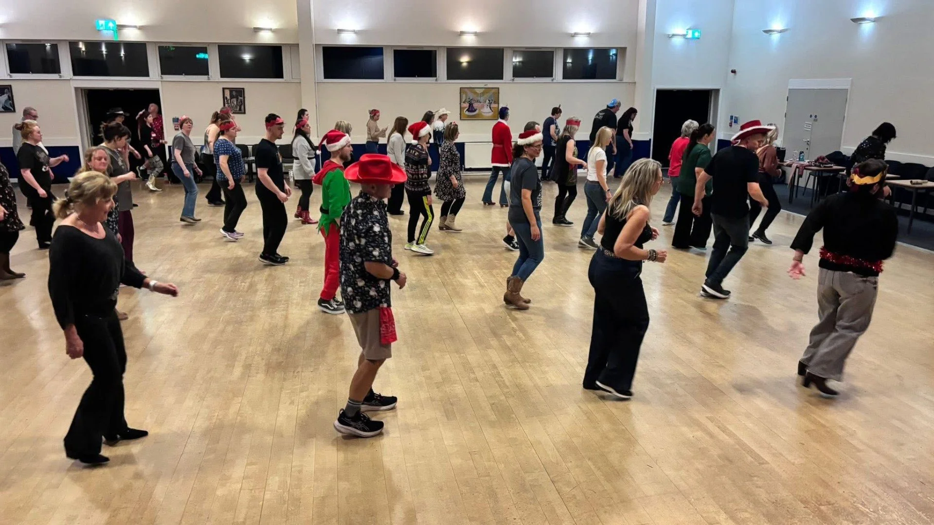 Group of people dancing in a hall with Christmas hats and festive clothing, celebrating at a holiday event.
