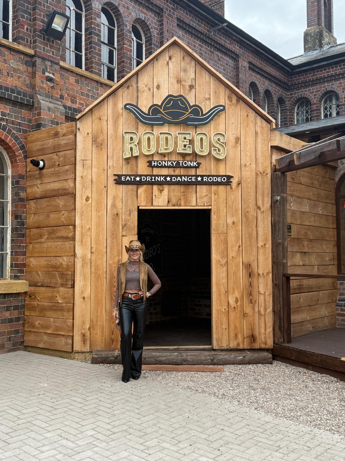 Woman standing in front of a wooden entrance to a rodeo event named 'Rodeos' with cowboy hat, leather pants, and long blonde hair, with brick building in the background.