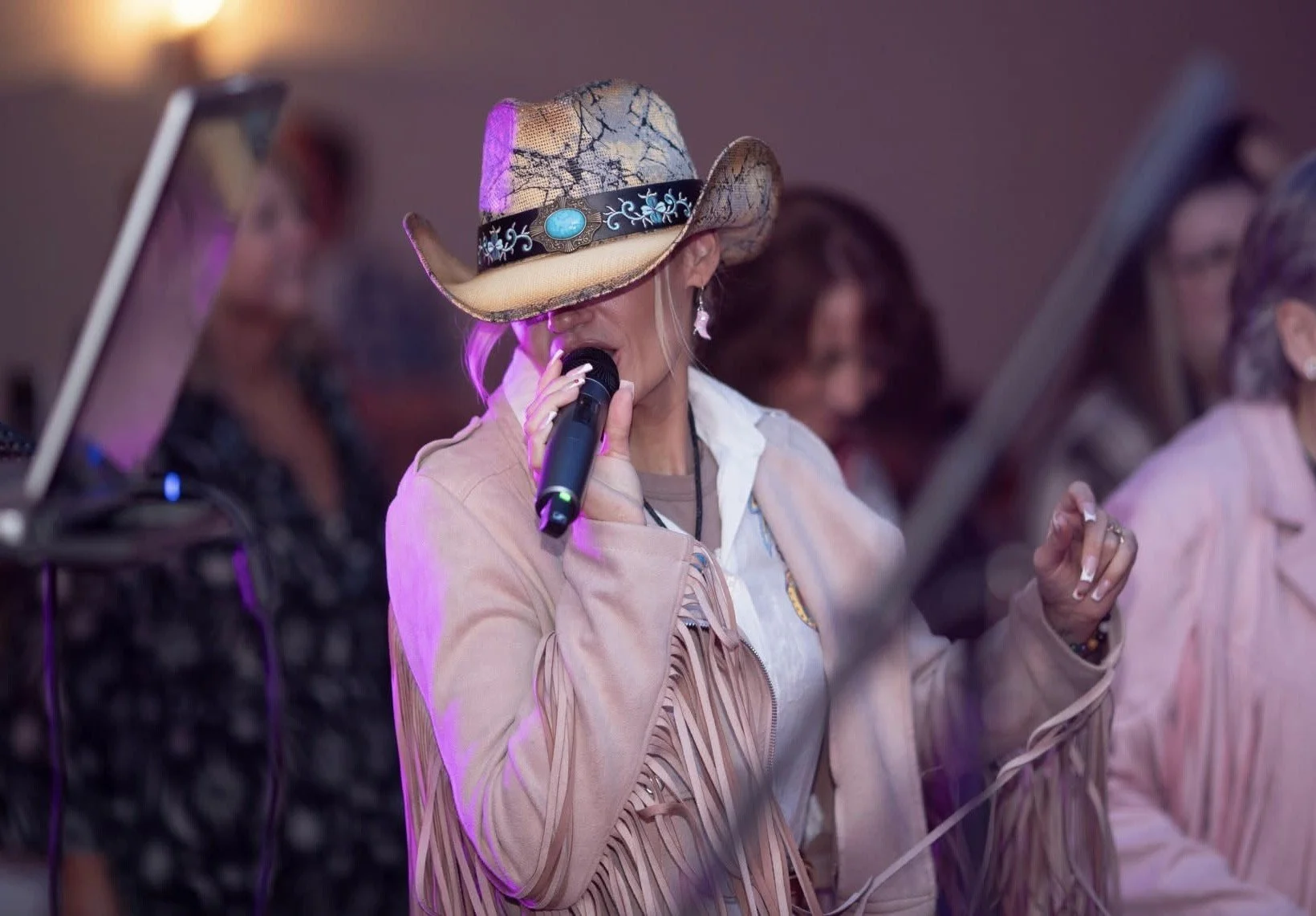 A woman wearing a fringed jacket and a cowboy hat singing into a microphone at a karaoke or music event, with others in the background.