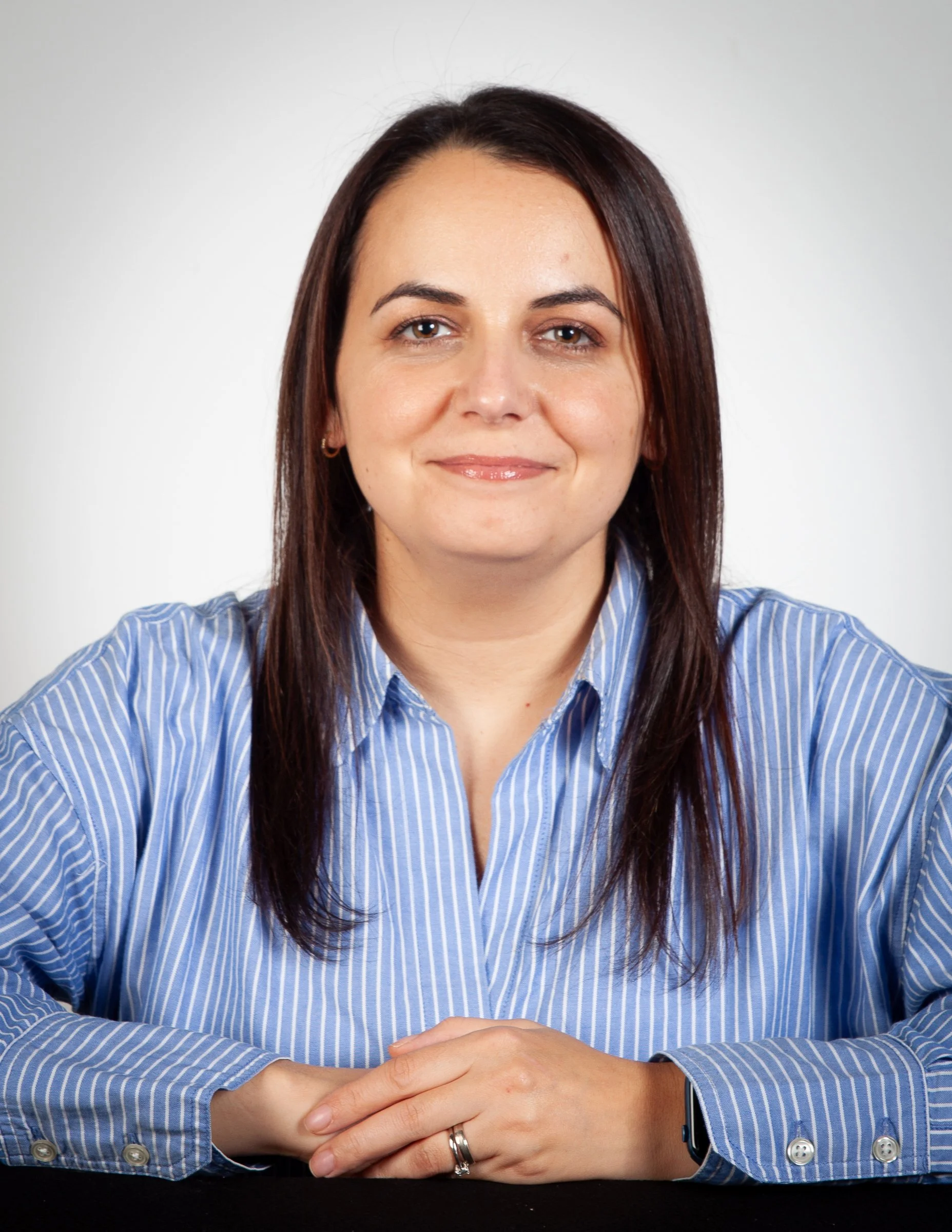 Portrait of a woman with shoulder-length dark brown hair, wearing a blue and white striped button-up shirt, sitting at a table with a white background.