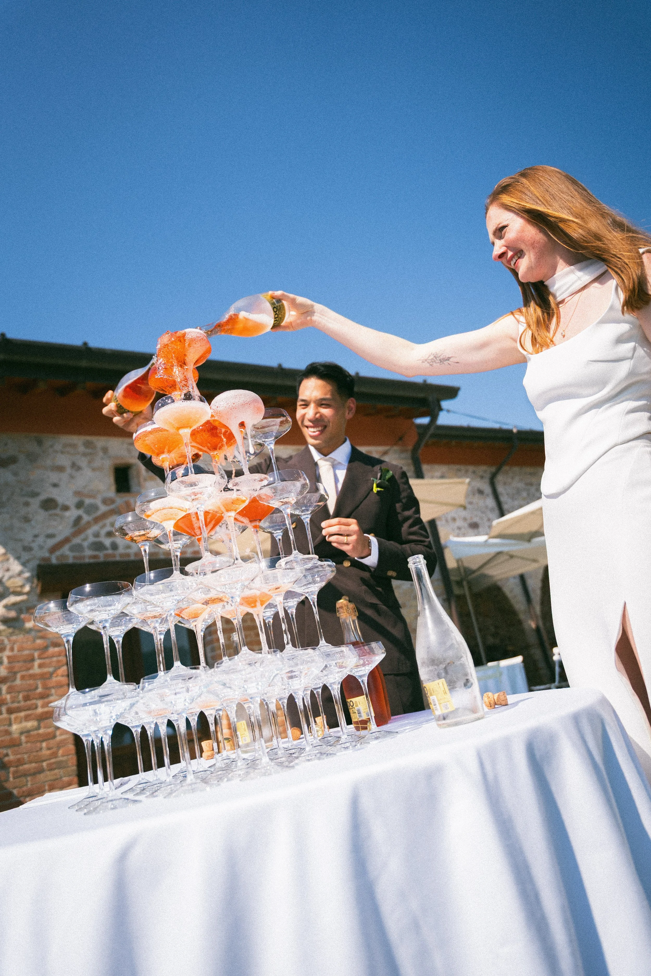 Una scena di festa con una donna con cappello di pelle rosa che versa champagne in un tower di bicchieri di vetro. Sul tavolo c'è una bottiglia di champagne e un recipiente di vetro. Sullo sfondo un uomo in abito e una parete di mattoni