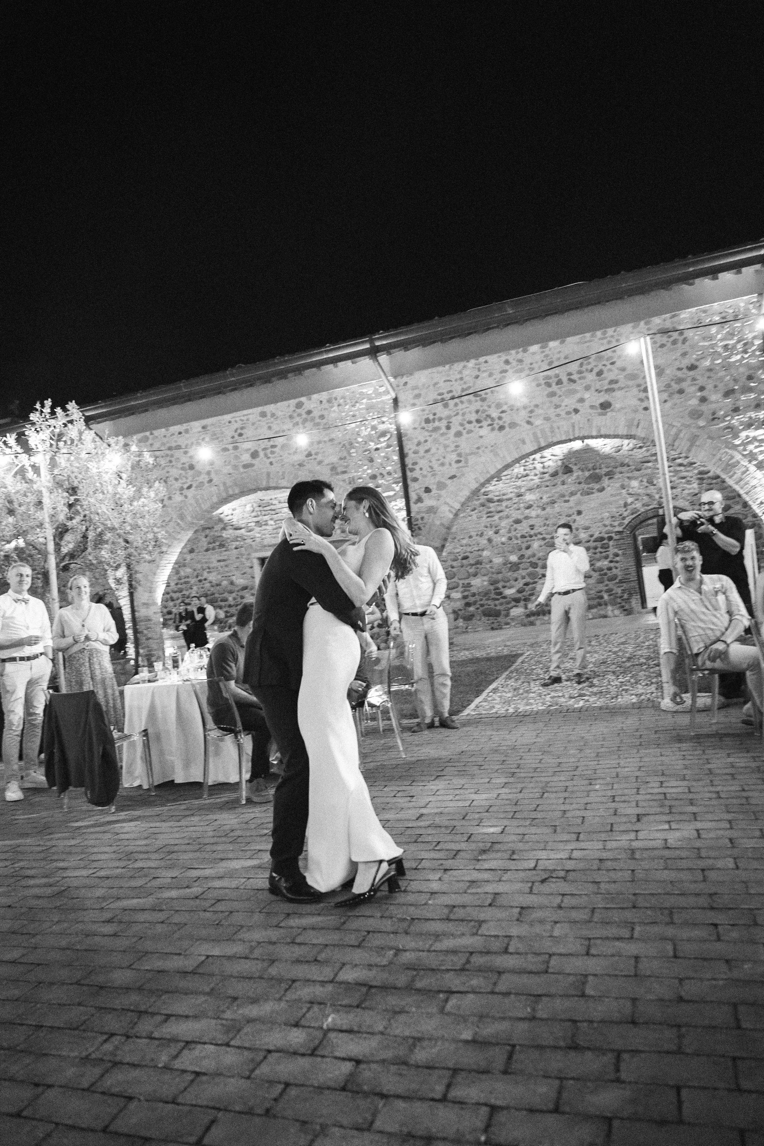 Un uomo e una donna ballano insieme in un'atmosfera romantica all'aperto di sera, con persone che assistono e un ponte in pietra sullo sfondo.