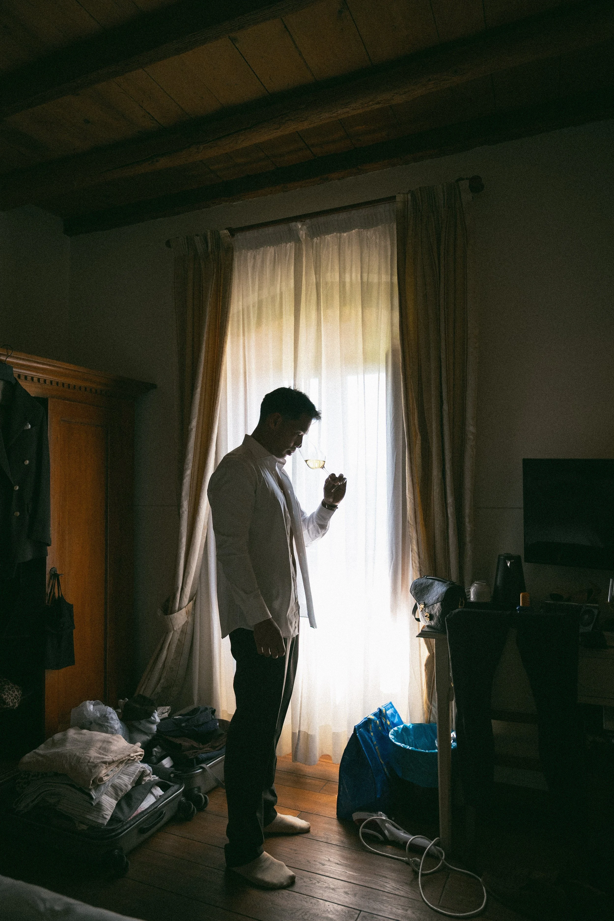 Un uomo con abiti eleganti sta riflettendo con approccio serio in una stanza con luci soffuse, vicino a una finestra con tende leggere. La stanza ha un soffitto in legno e mobili in legno, con vestiti e oggetti sparsi sul pavimento e sui mobili.