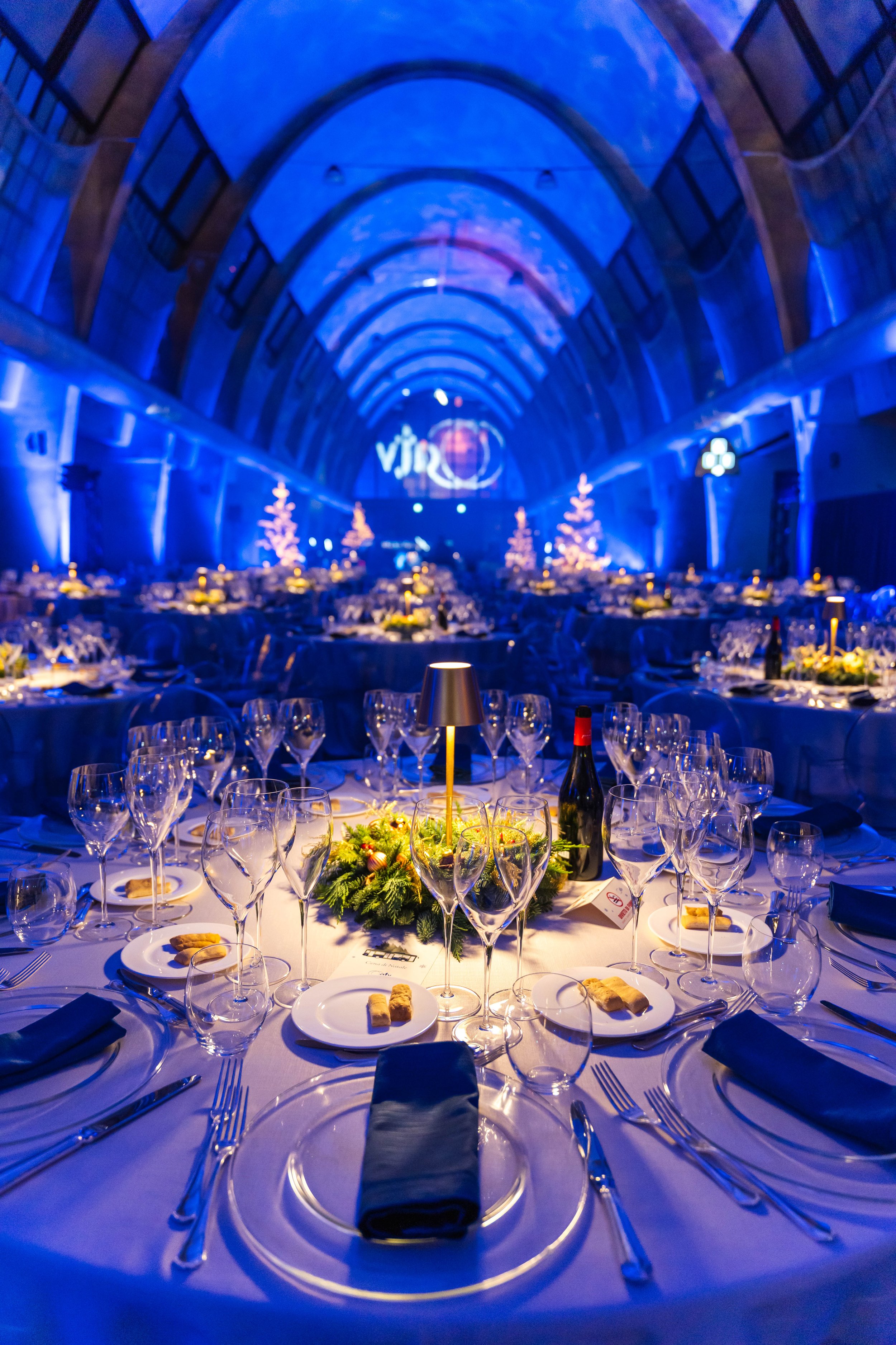 Sala da banchetto natalizia con luci blu, tavoli decorati con candelabri e centrotavola verdi, presenta un'atmosfera festiva.