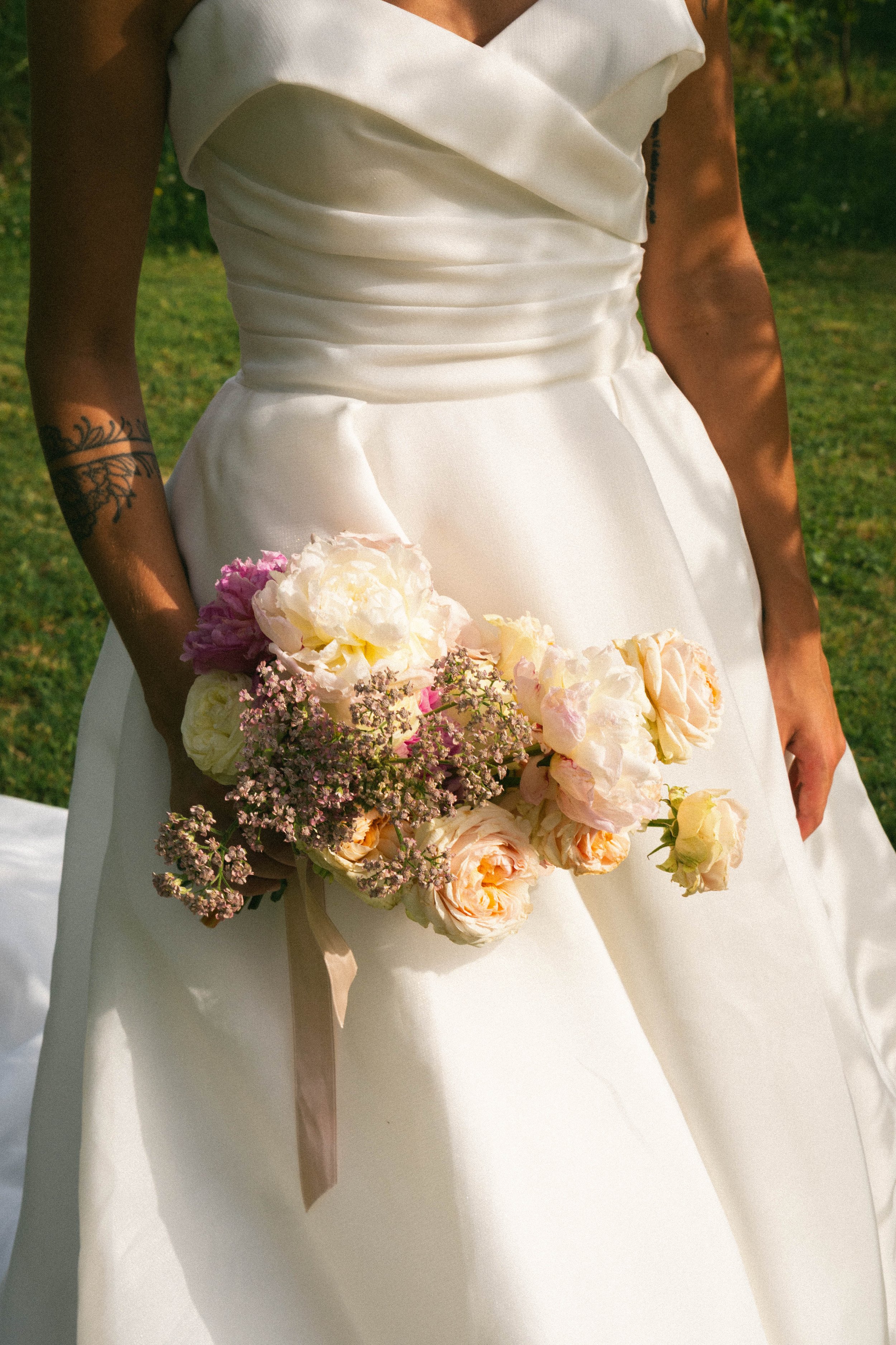 Una donna indossa un abito da sposa bianco e tiene un bouquet di fiori, in una scena all'aperto.