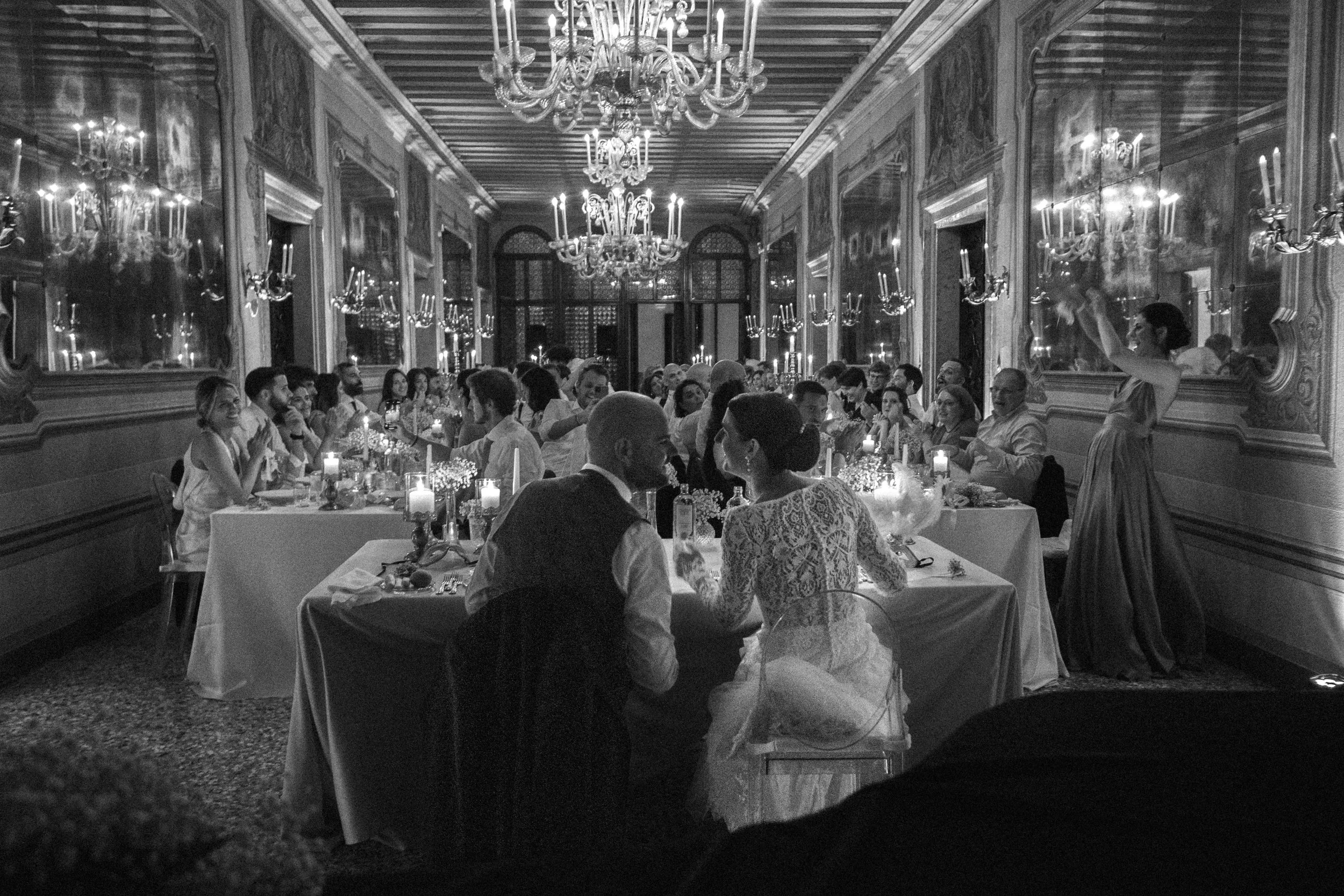 Una grande sala elegante con soffitto in legno e lampadari di cristallo, piena di persone sedute a tavoli con candele, in occasione di un evento o cena formale, con decorazioni floreali e specchi alle pareti.