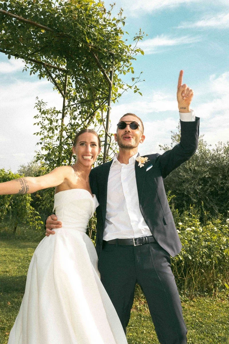 Una coppia di sposi felici si scatta una foto all'aperto durante il loro matrimonio; la donna indossa un abito bianco e il uomo un abito scuro con occhiali da sole.