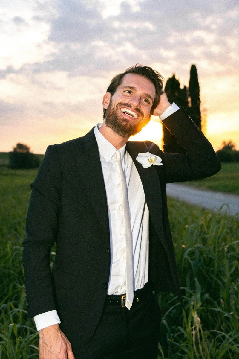 Uomo sorridente con abito nero e camicia bianca in un campo durante il tramonto.