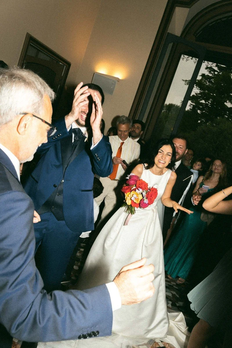 Una coppia di sposi felice, la donna con un bouquet di fiori, circondati da amici e parenti durante un matrimonio in una sala elegante con grandi finestre vista sul giardino