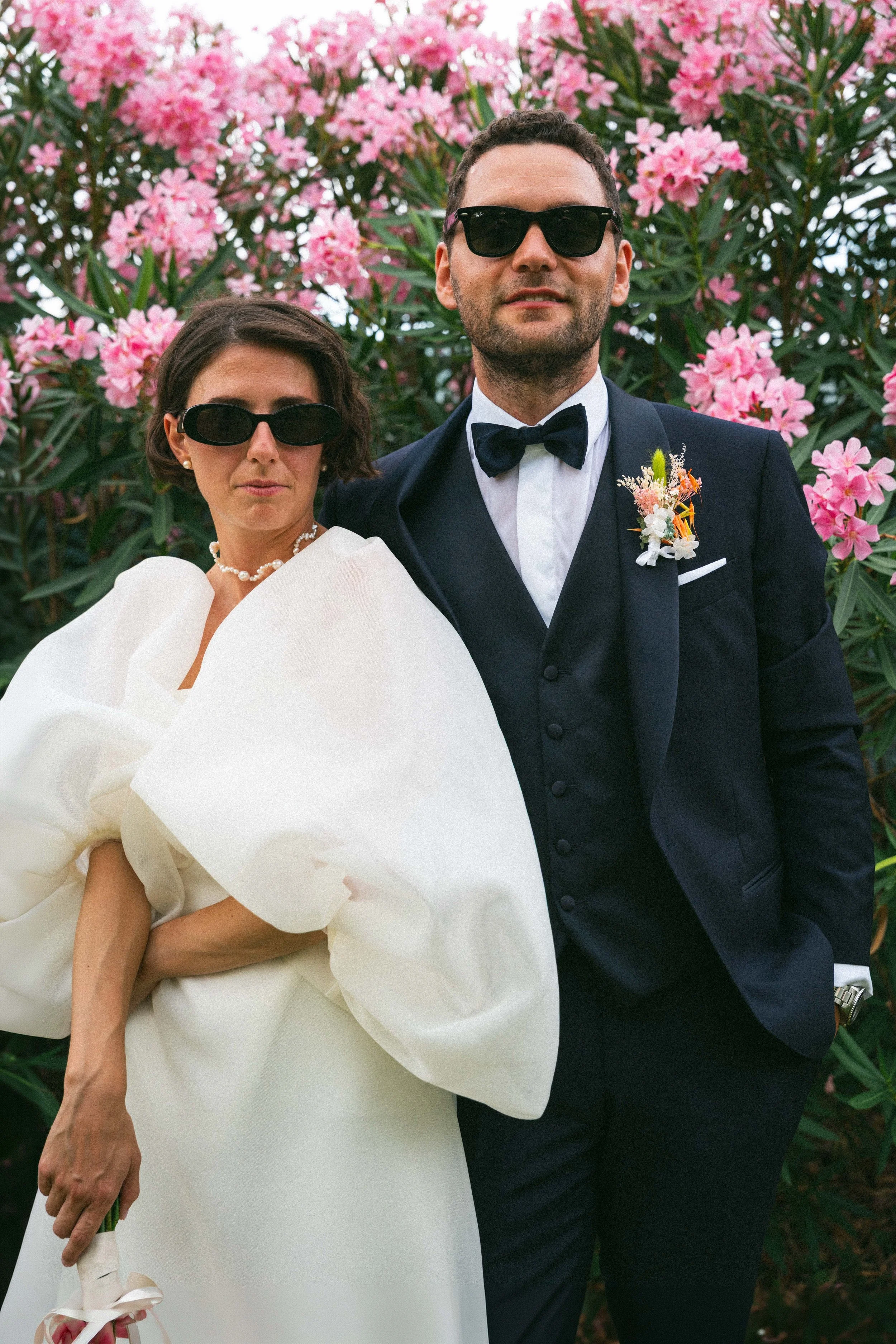 Una coppia elegante, l'uomo con un completo nero con fiocco e boutonnière di fiori e la donna con un abito bianco con ampie maniche e collana di perle, indossano entrambe occhiali da sole scuri e sono in piedi di fronte a bshere di fiori rosa.