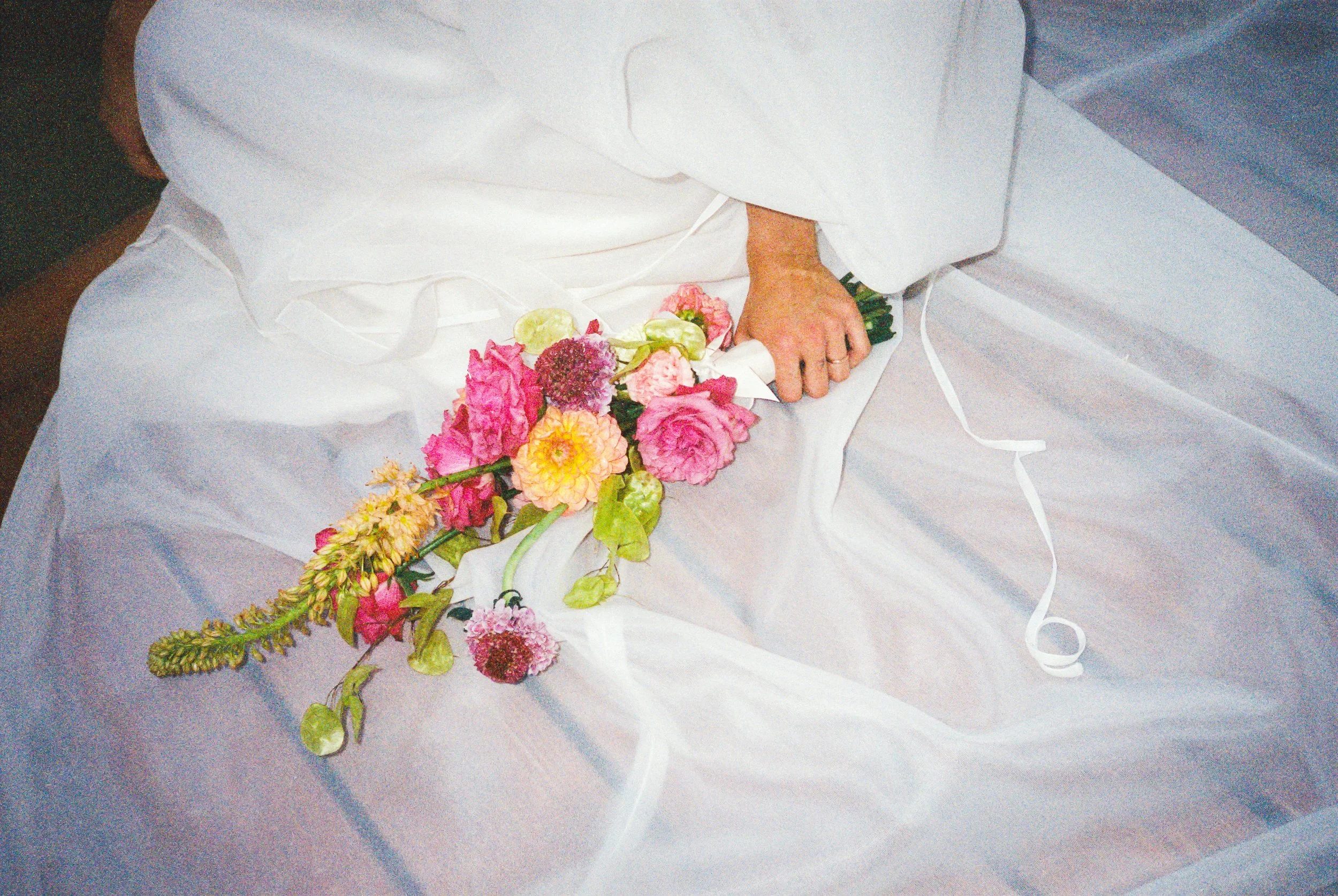 Una mano, probabilmente di una donna, tiene un mazzo di fiori di diversi colori e tipi, appoggiato su un tessuto bianco.