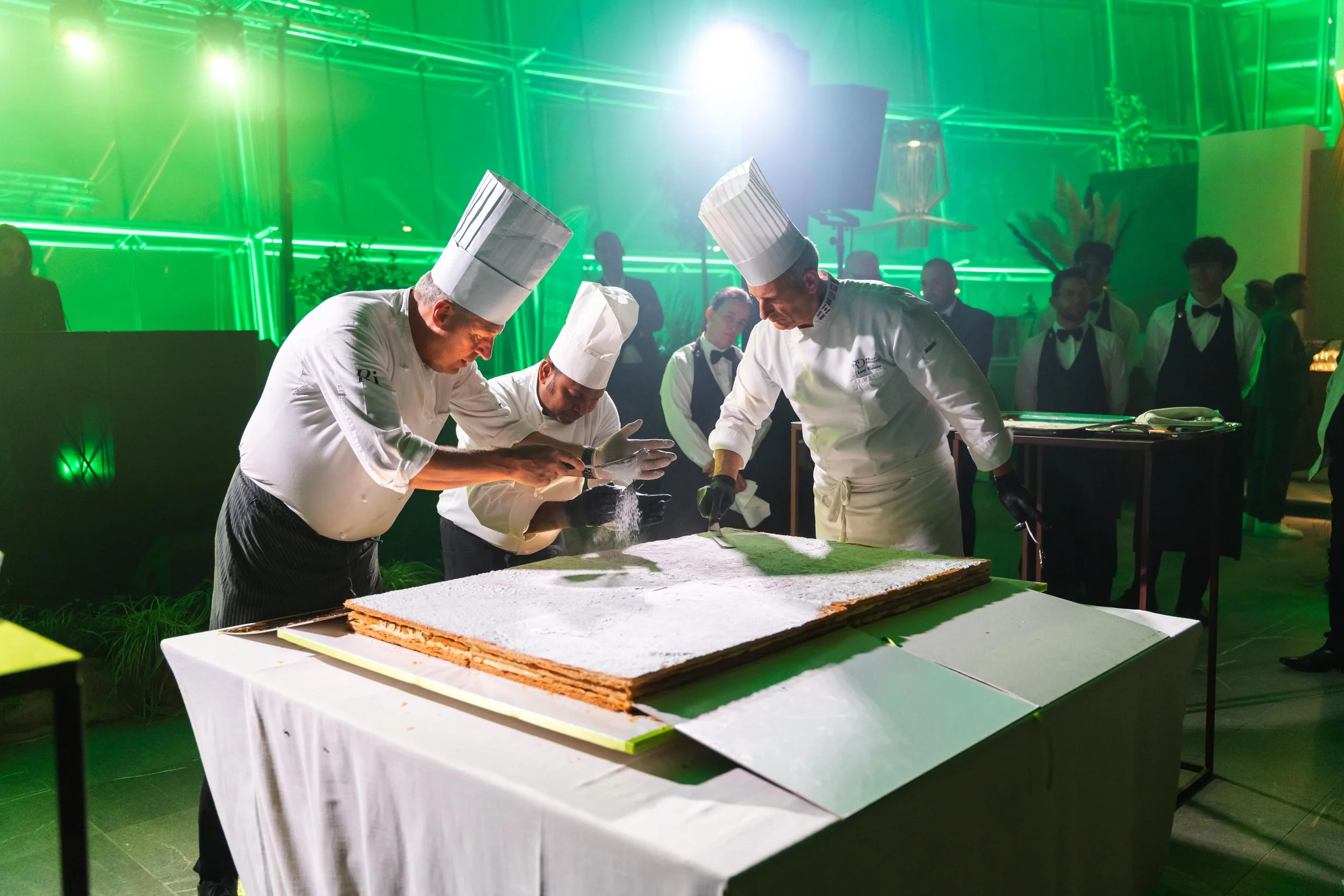 Chef decorando un grande dolce con zucchero a velo in un evento con luci verdi, con altri chef e camerieri che assistono.