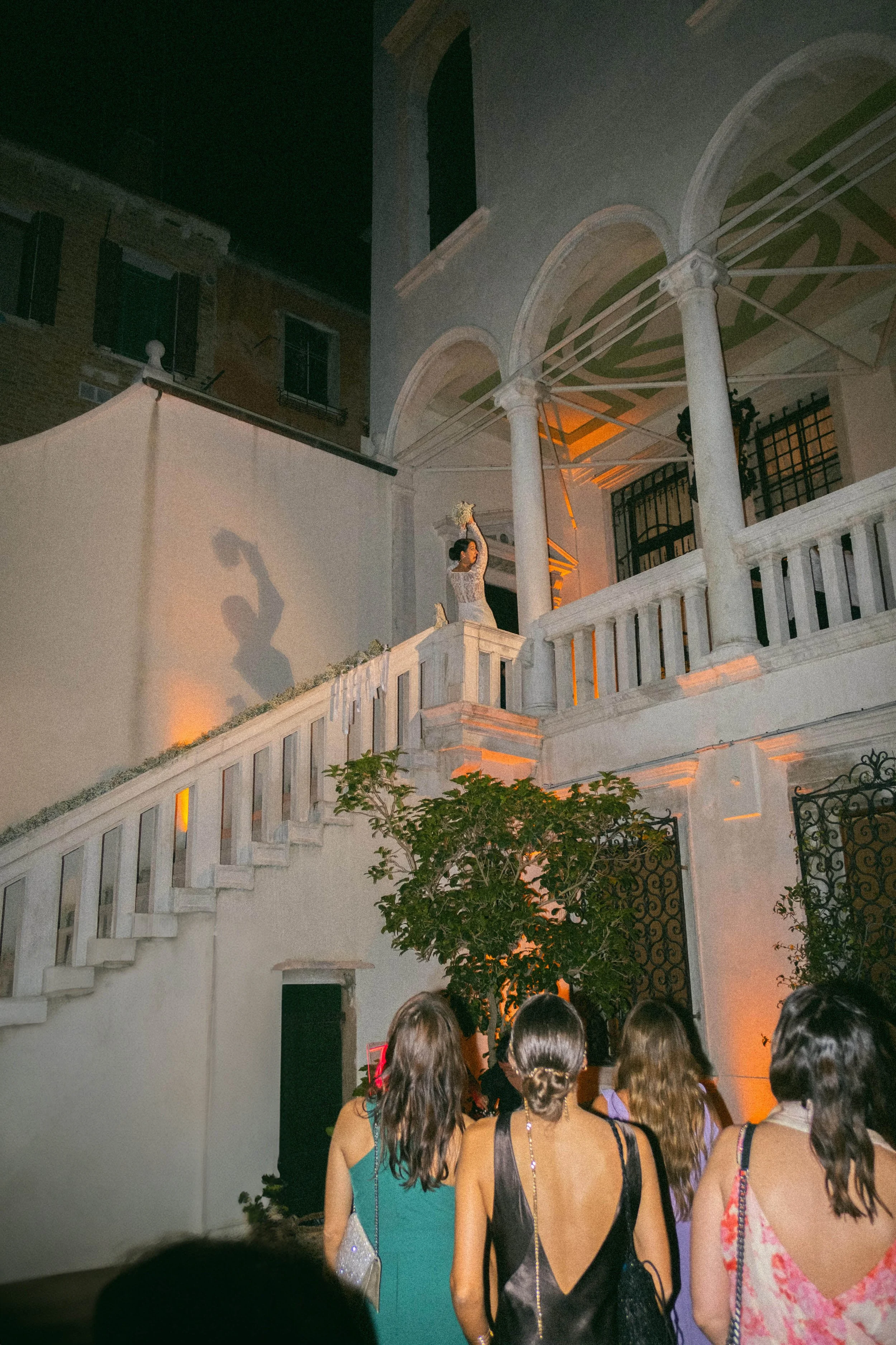 Folla di persone che guarda uno spettacolo all'esterno durante la sera, con una struttura architettonica bianca, colonne e un muro con posa affacciata di case in stile antico