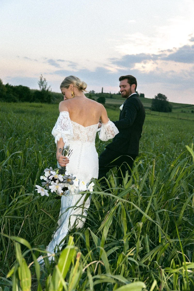 Una coppia di sposi in abiti da matrimonio si tiene per mano in un campo di erba verde, con un cielo nuvoloso sullo sfondo.