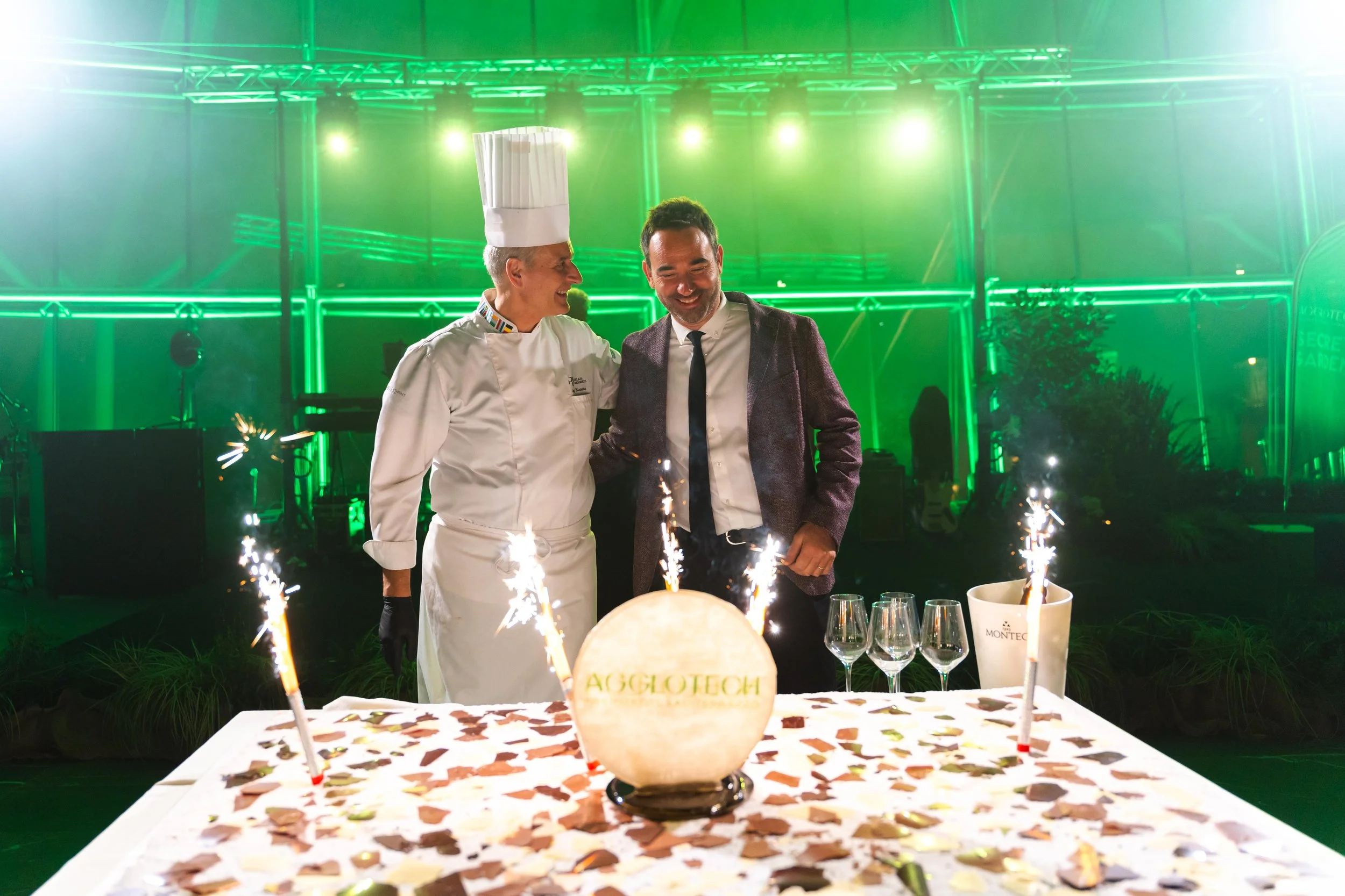 Chef e uomo in abito elegante sorridenti e vicini davanti a una torta decorata con scintille, su un tavolo con bicchieri di vino, in un ambiente festoso con luci verdi sullo sfondo.