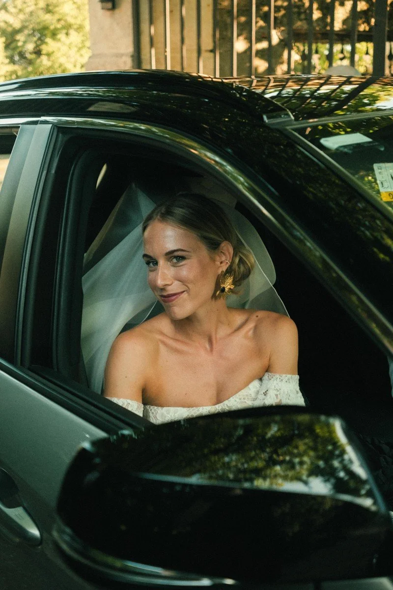 Una donna in abito da sposa seduta in una macchina, sorridente e con un velo, con sfondo di una residenza e alberi