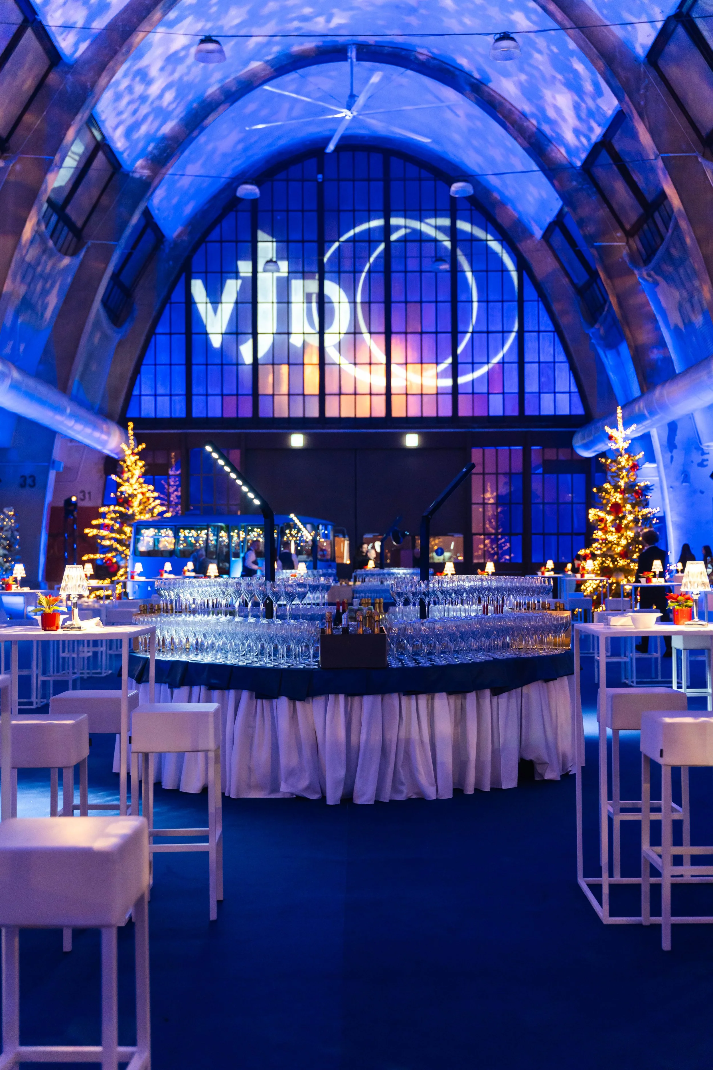 Interno di un locale elegante con decorazioni natalizie, due alberi di Natale illuminati, tavoli con candelabri e bicchieri, e un grande soffitto a volta con proiezione di luci e il logo Vitro.