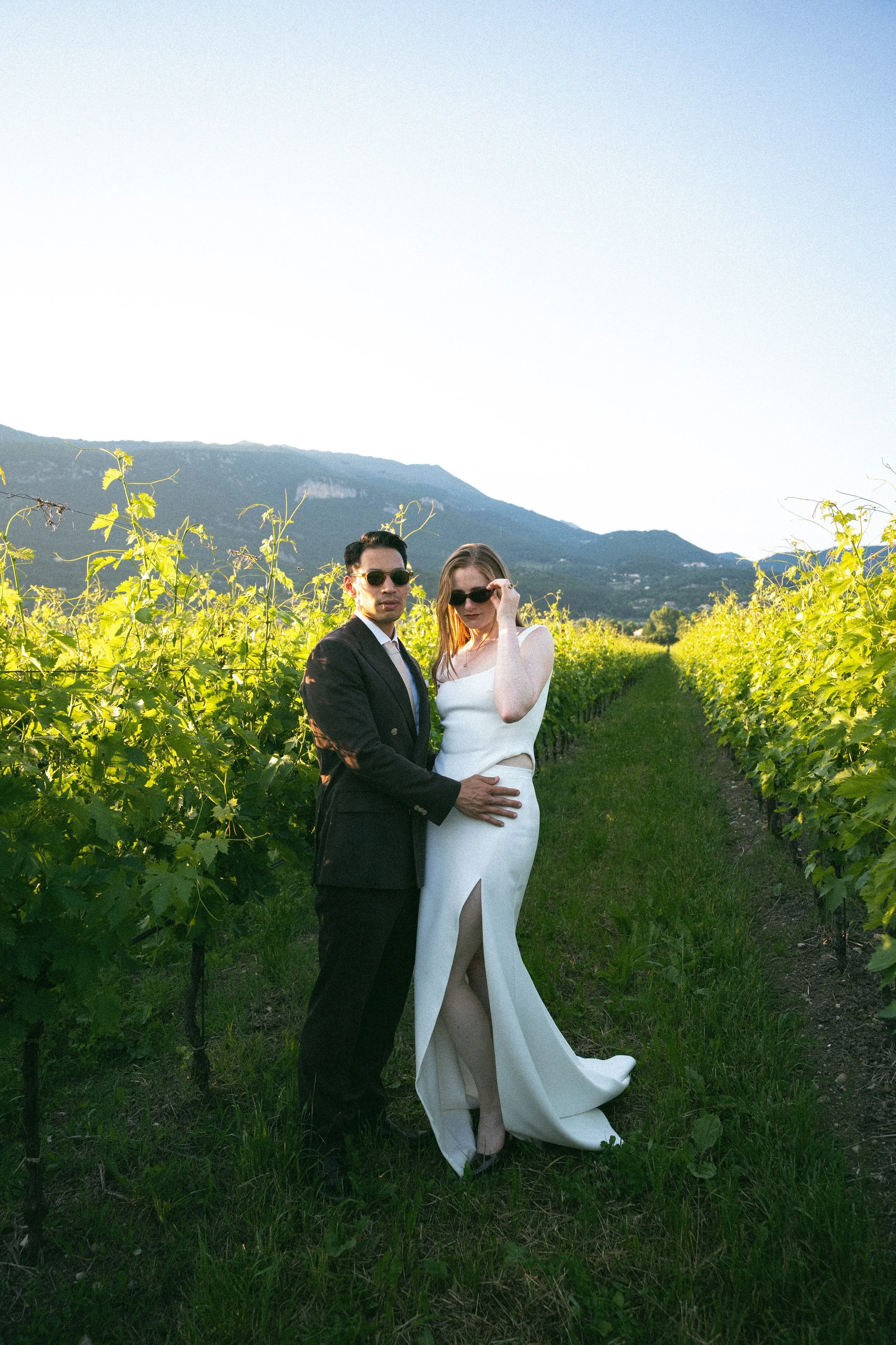 Una coppia elegante in abiti da sera in una vigna con montagne sullo sfondo sotto un cielo chiaro.