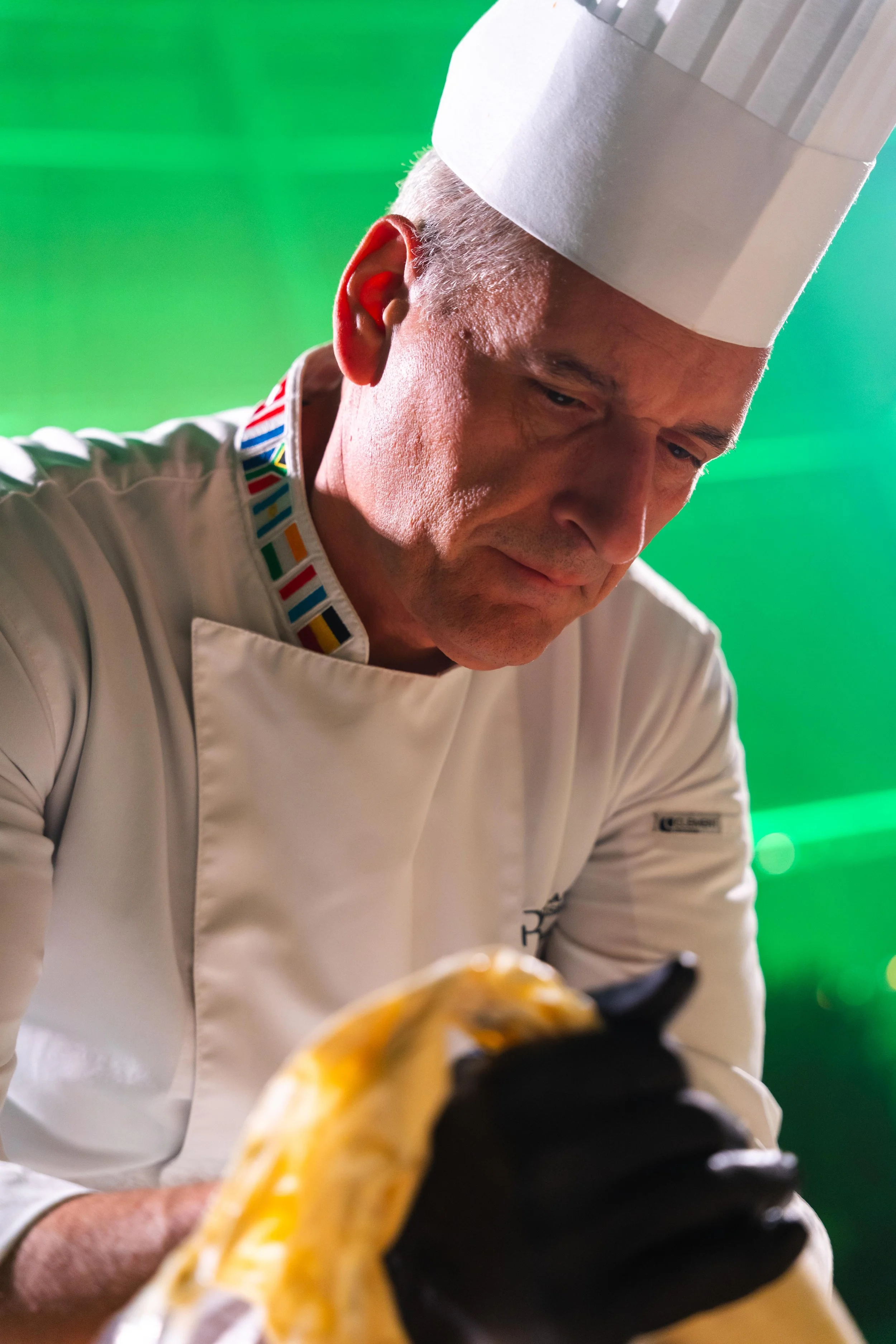 Chef in divisa bianca con cappello da cuoco, che lavora con attenzione con un panno giallo e nero in un ambiente con luce verde.