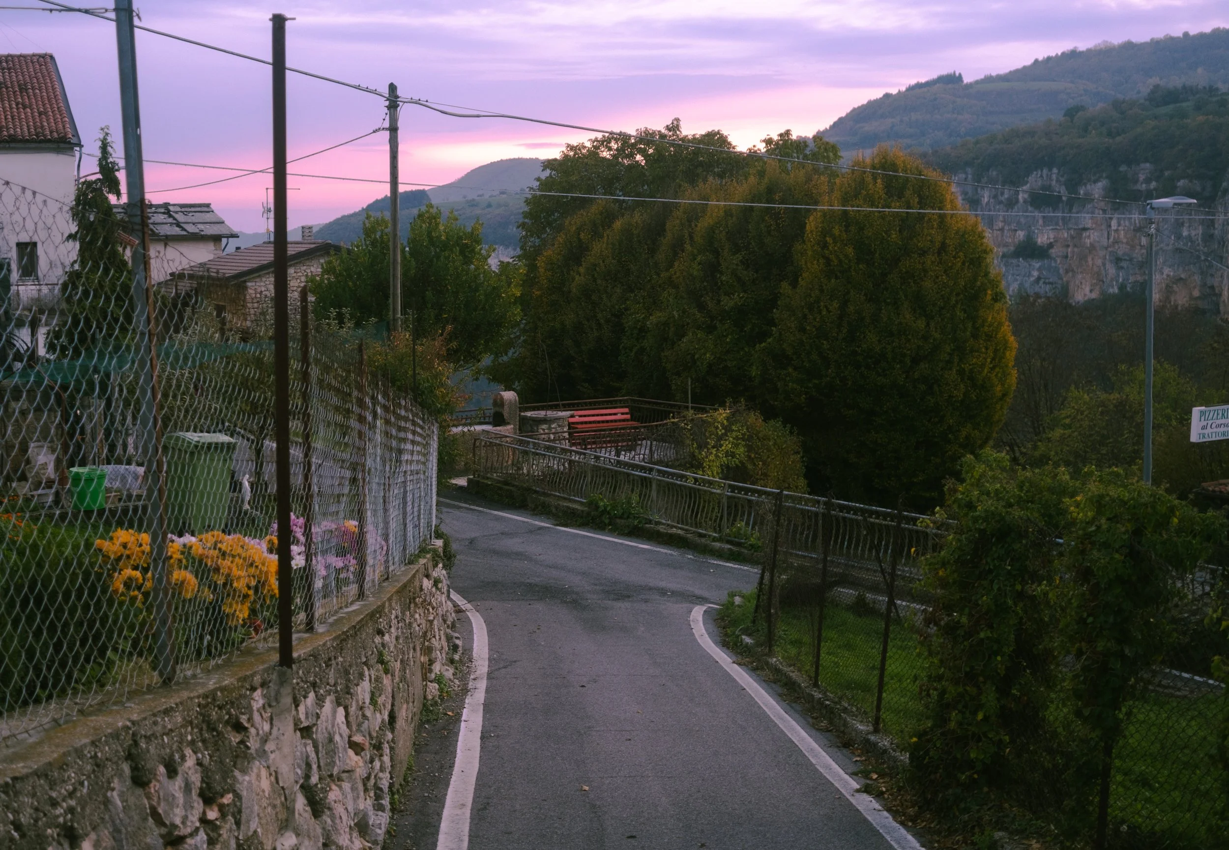 Stradina di montagna al tramonto con case, fiori, alberi e montagne sullo sfondo.
