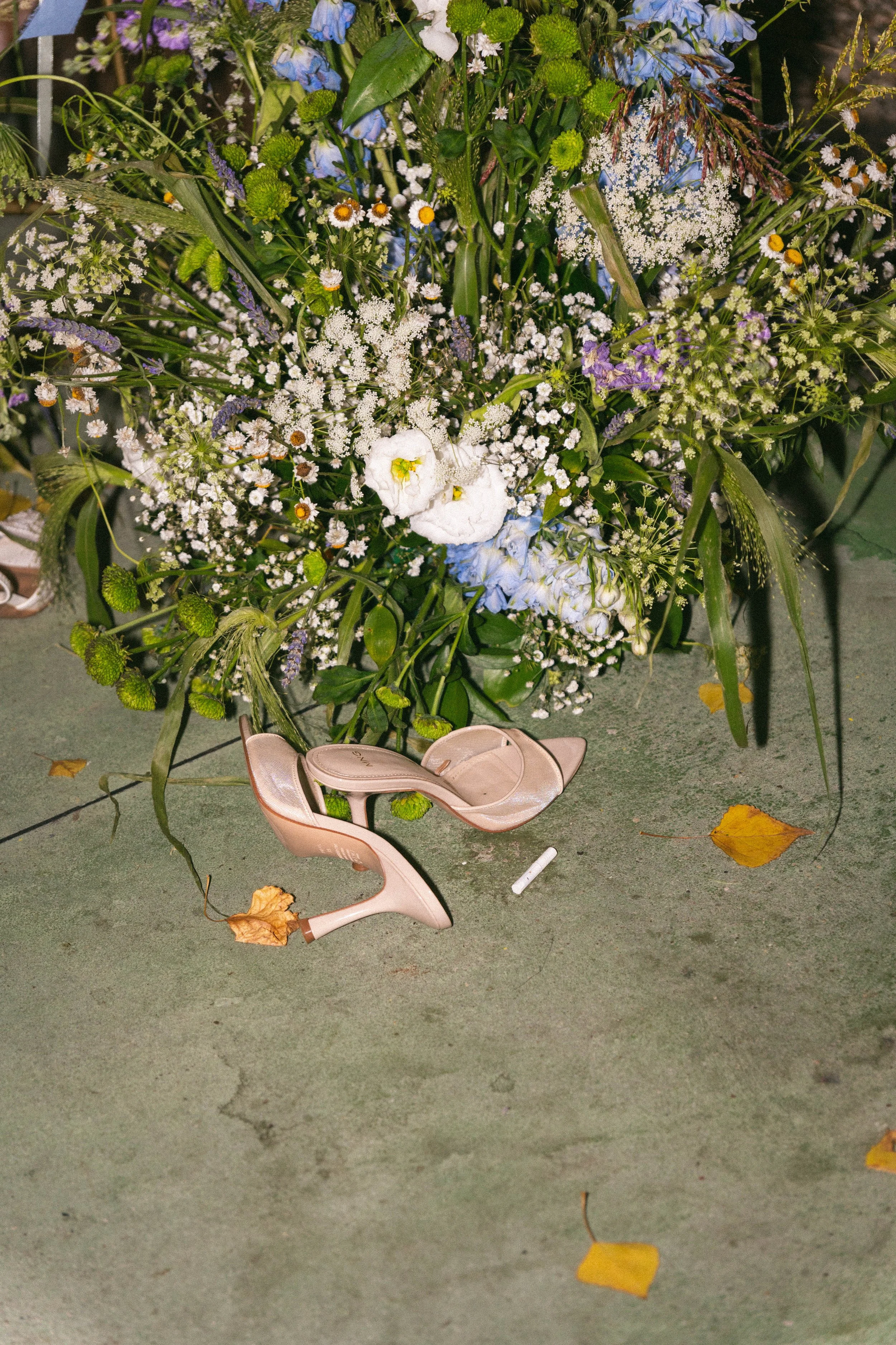 Un grande mazzo di fiori colorati, inclusi fiori bianchi, blu, viola e gialli, poggiato su un centro di tavolo. Davanti si trovano un paio di scarpe rosa, un pennarello bianco e alcune foglie secche sparse sul pavimento.