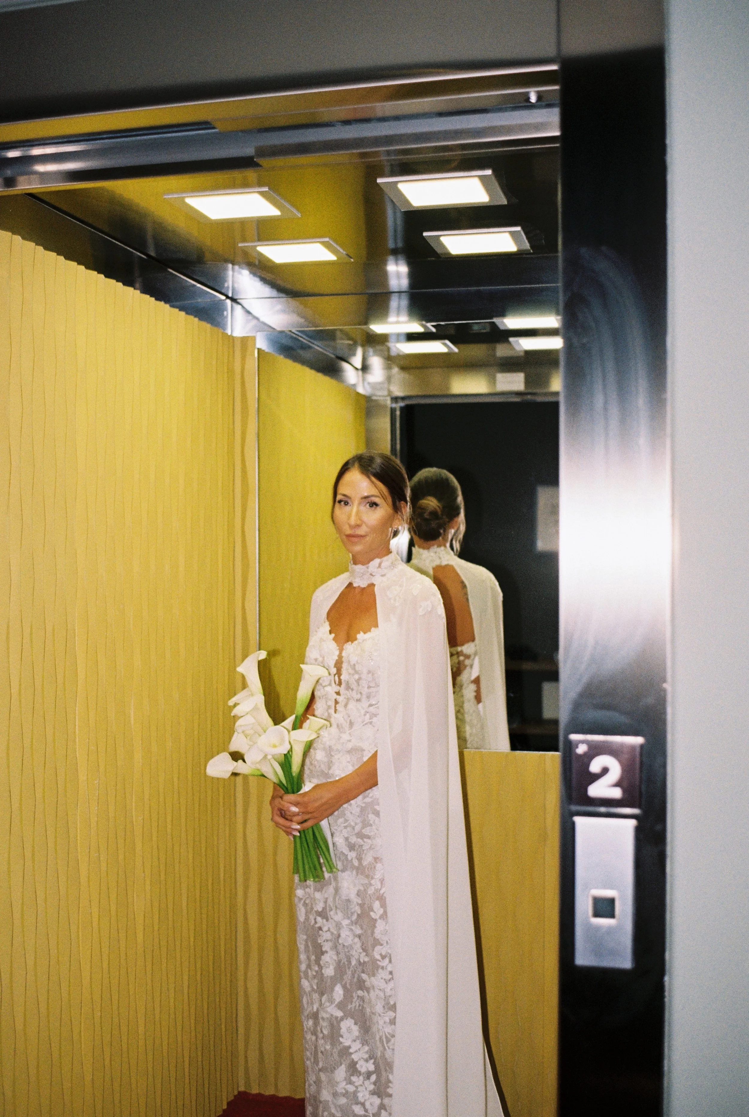 Una donna con un abito bianco in un ascensore, con uno sfondo di specchio e pareti gialle, tiene un mazzo di calle bianche e guarda verso la fotocamera.
