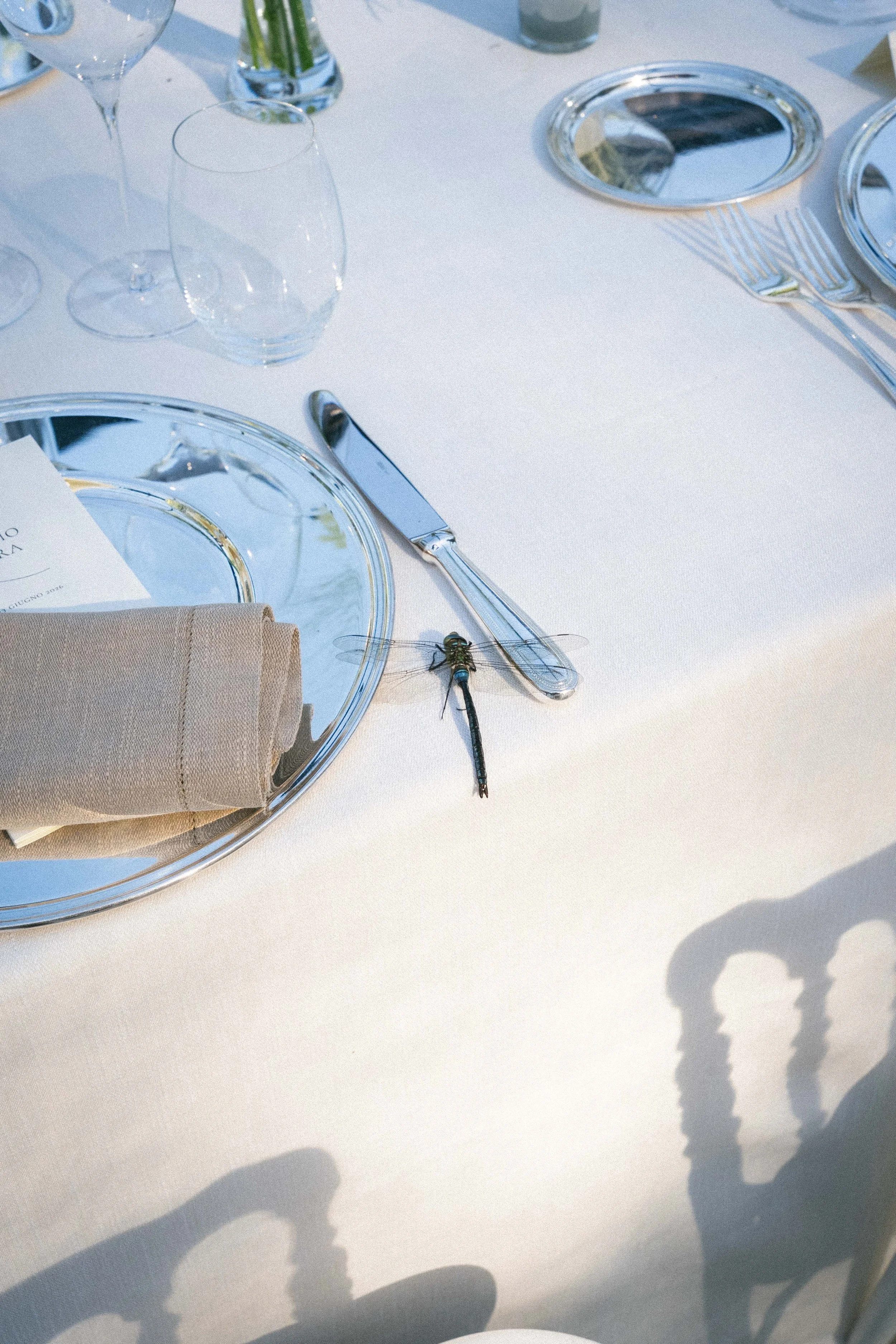 Tavola apparecchiata con piatti di vetro, coltelli, forchette e bicchieri, con un tovagliolo di stoffa e un menu, decorazioni floreali, luce naturale, ombre delle mani o delle persone intorno alla tavola.