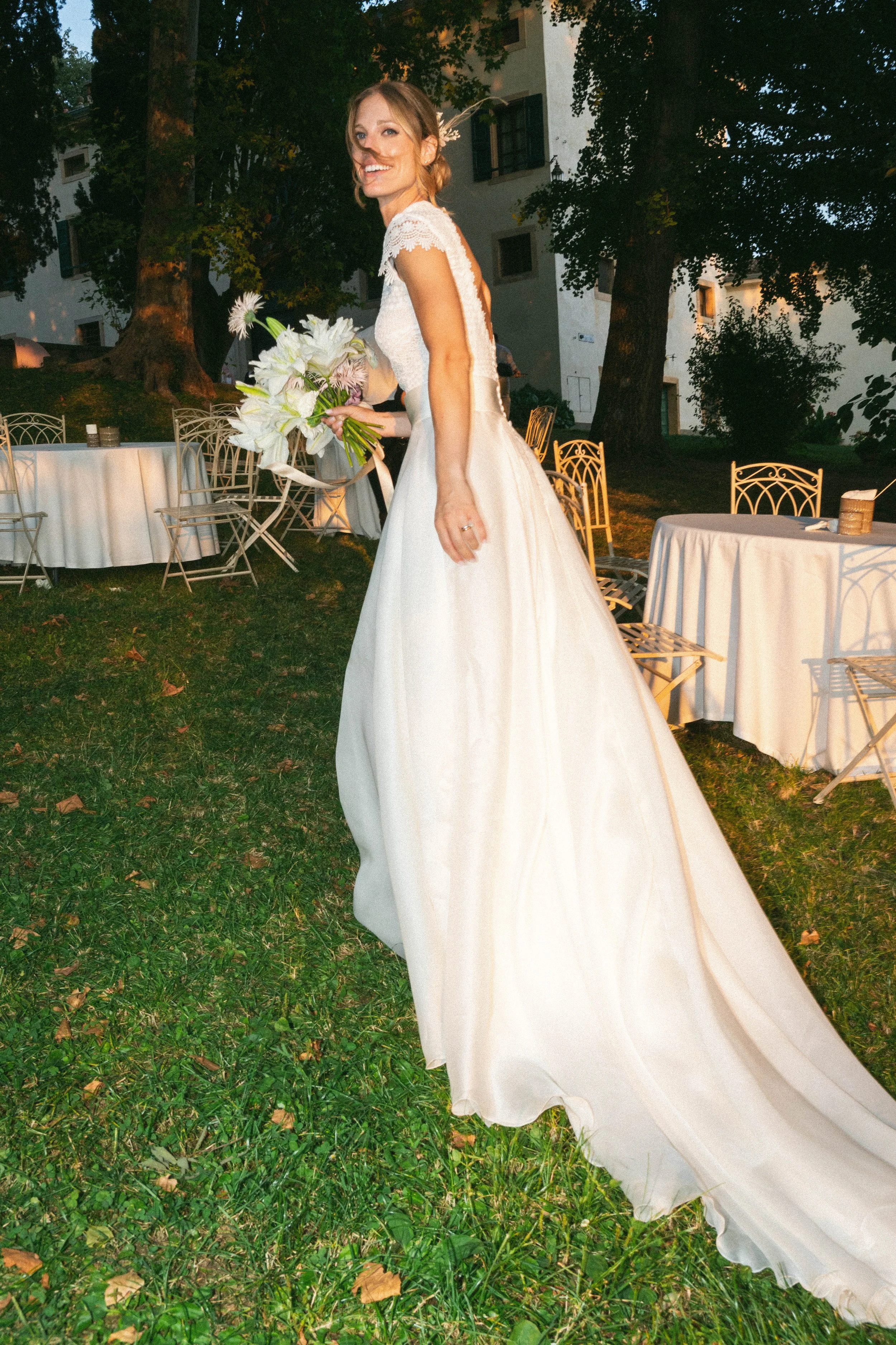 Una donna in abito da sposa sorride mentre tiene un mazzo di fiori, fotografata di sera all'aperto con tavoli e sedie in metallo bianco e grandi alberi sullo sfondo.