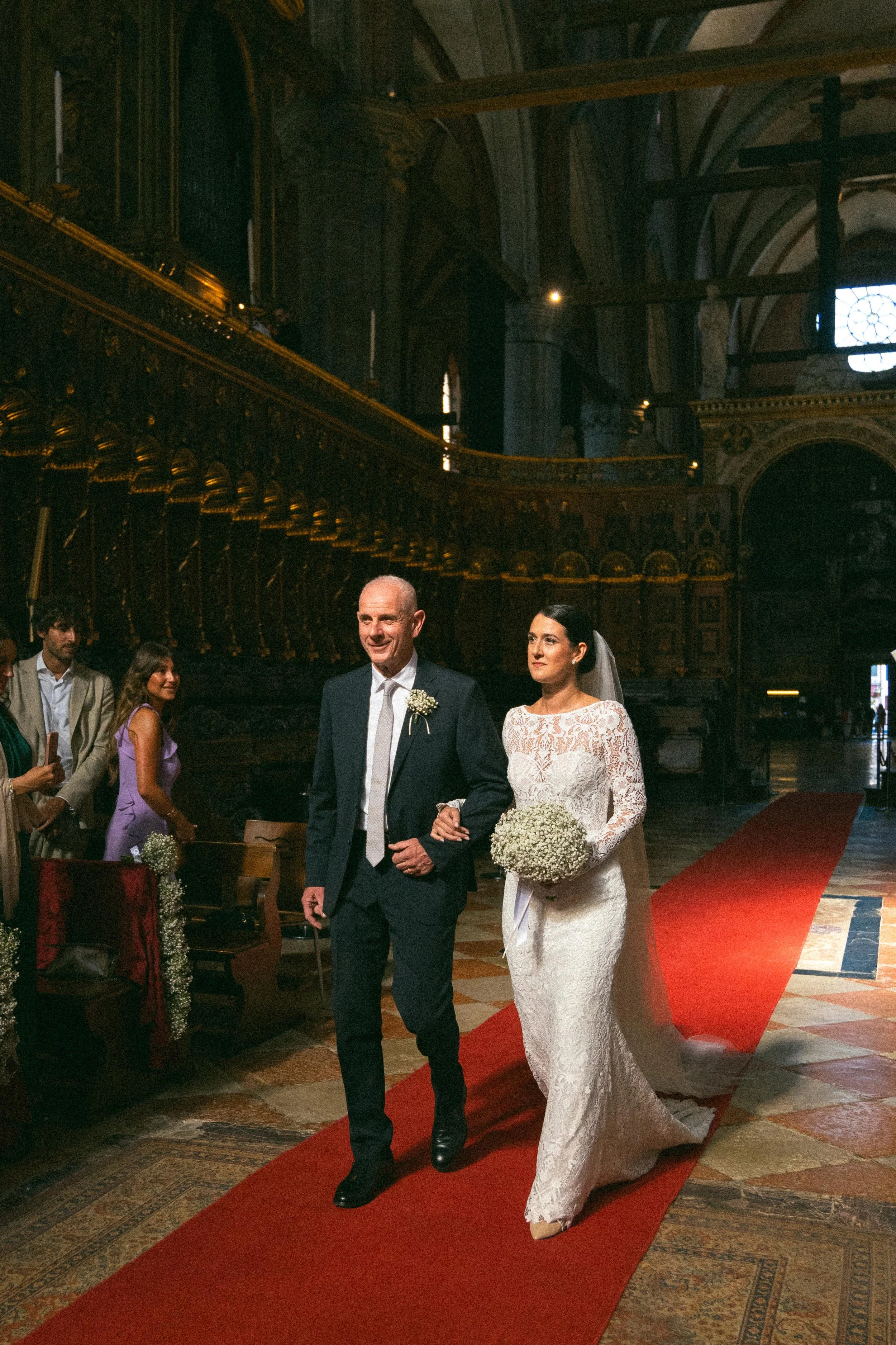 Una coppia si sposa in chiesa, lei indossa un abito da sposa bianco lungo con un bouquet di fiori bianchi, lui indossa un completo scuro. Sono felici mentre camminano lungo il tradizionale tappeto rosso, circondati da altri invitati.