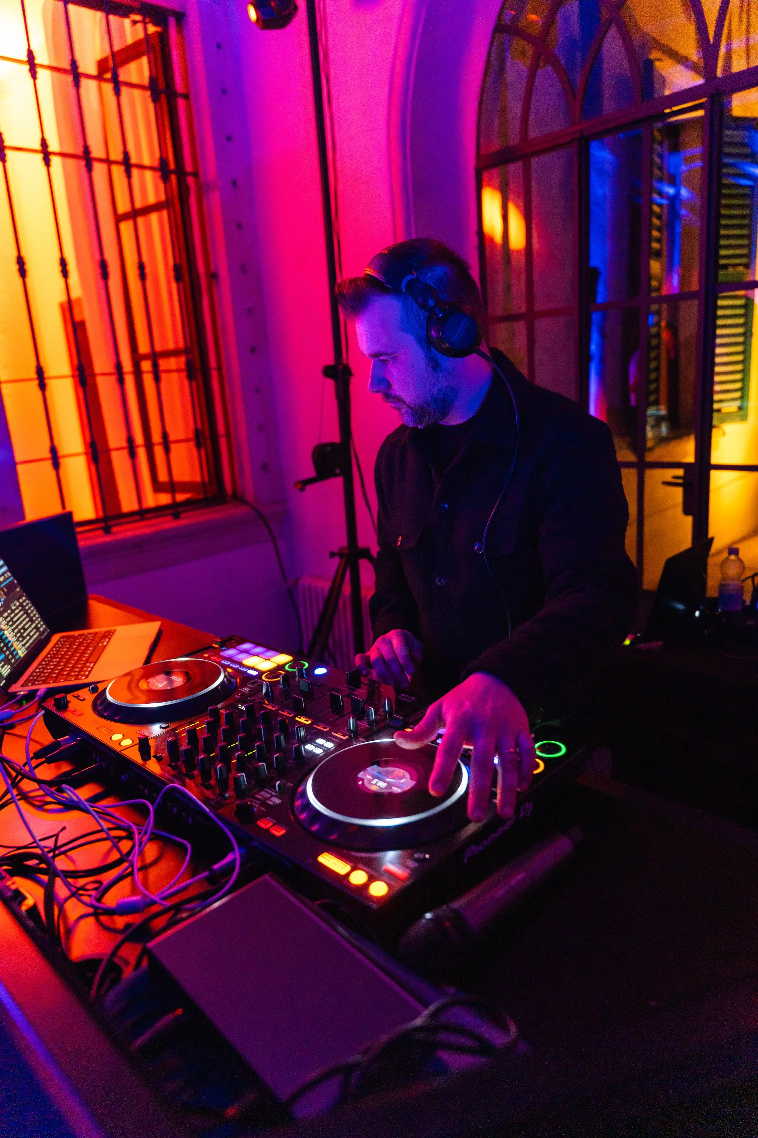 DJ che mixa con attrezzature da DJ, in un ambiente illuminato da luci colorate, con finestra e pareti in mattoni sullo sfondo.