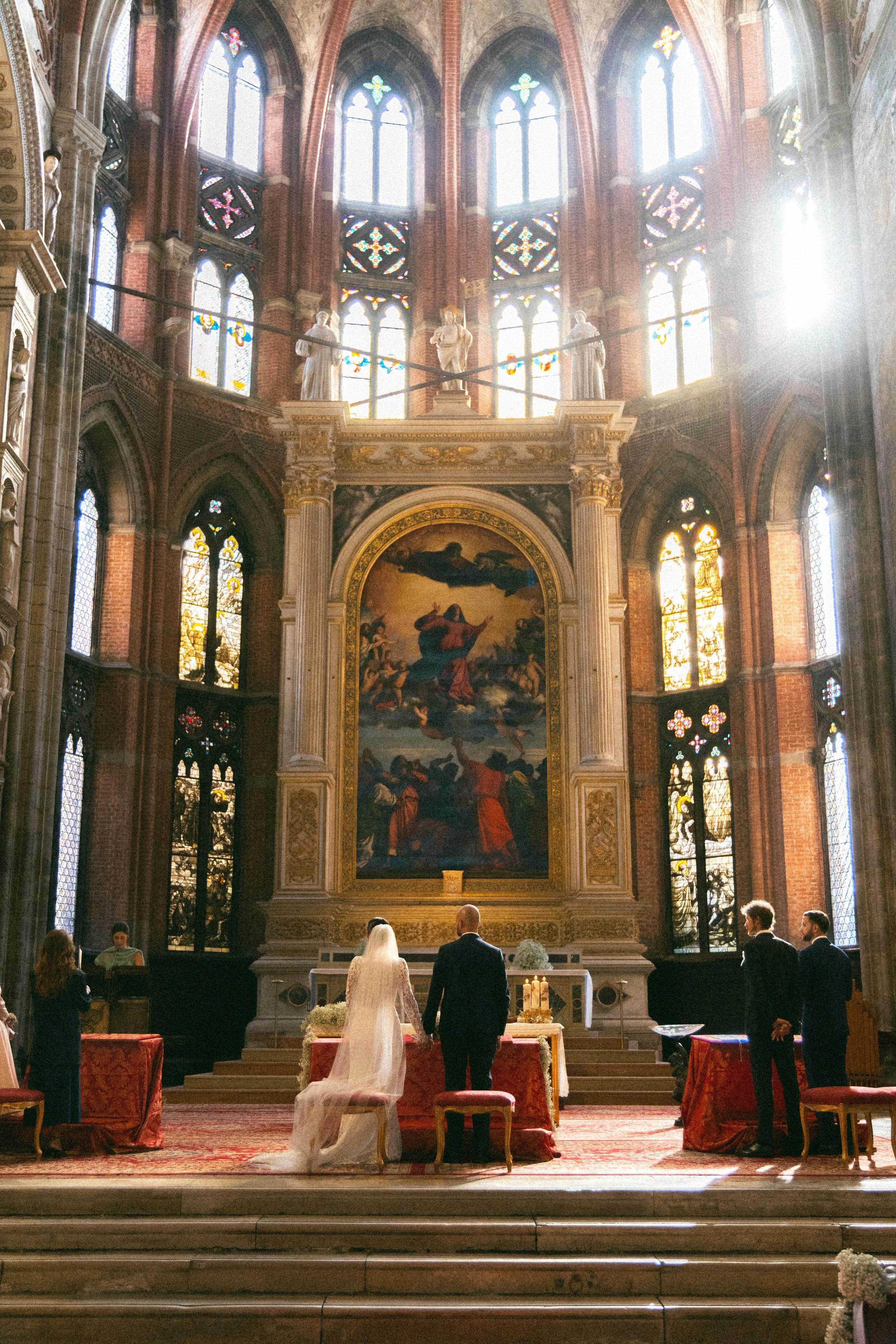 Cerimonia di matrimonio all'interno di una chiesa storica con vetrate colorate, statue e dipinti. Gli sposi sono davanti all'altare con ospiti seduti ai lati.
