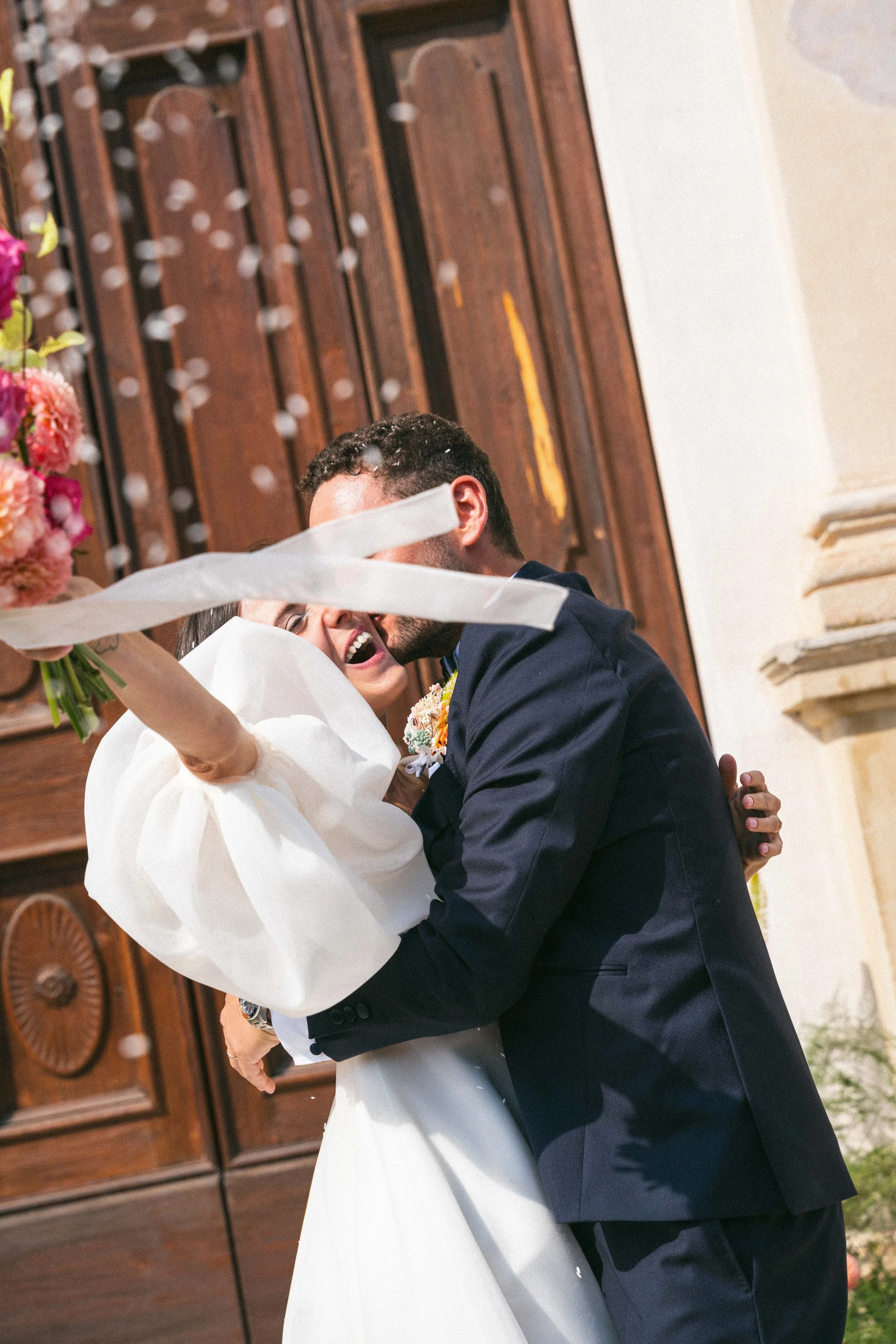 Una coppia di sposi si abbraccia e ride durante il loro matrimonio, con decorazioni floreali colorate sullo sfondo.