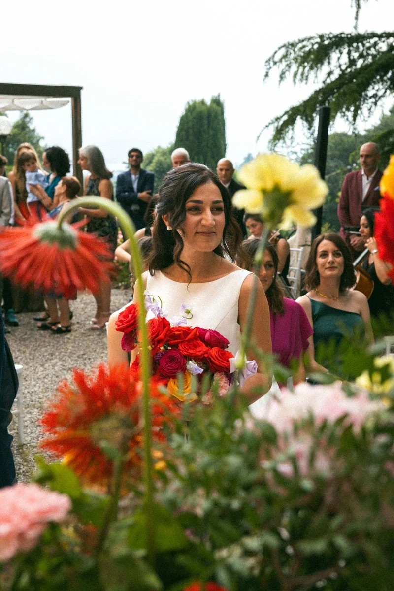 Una donna vestita elegantemente che tiene un mazzo di fiori colorati, circondata da altre persone in un evento all'aperto con decorazioni floreali.