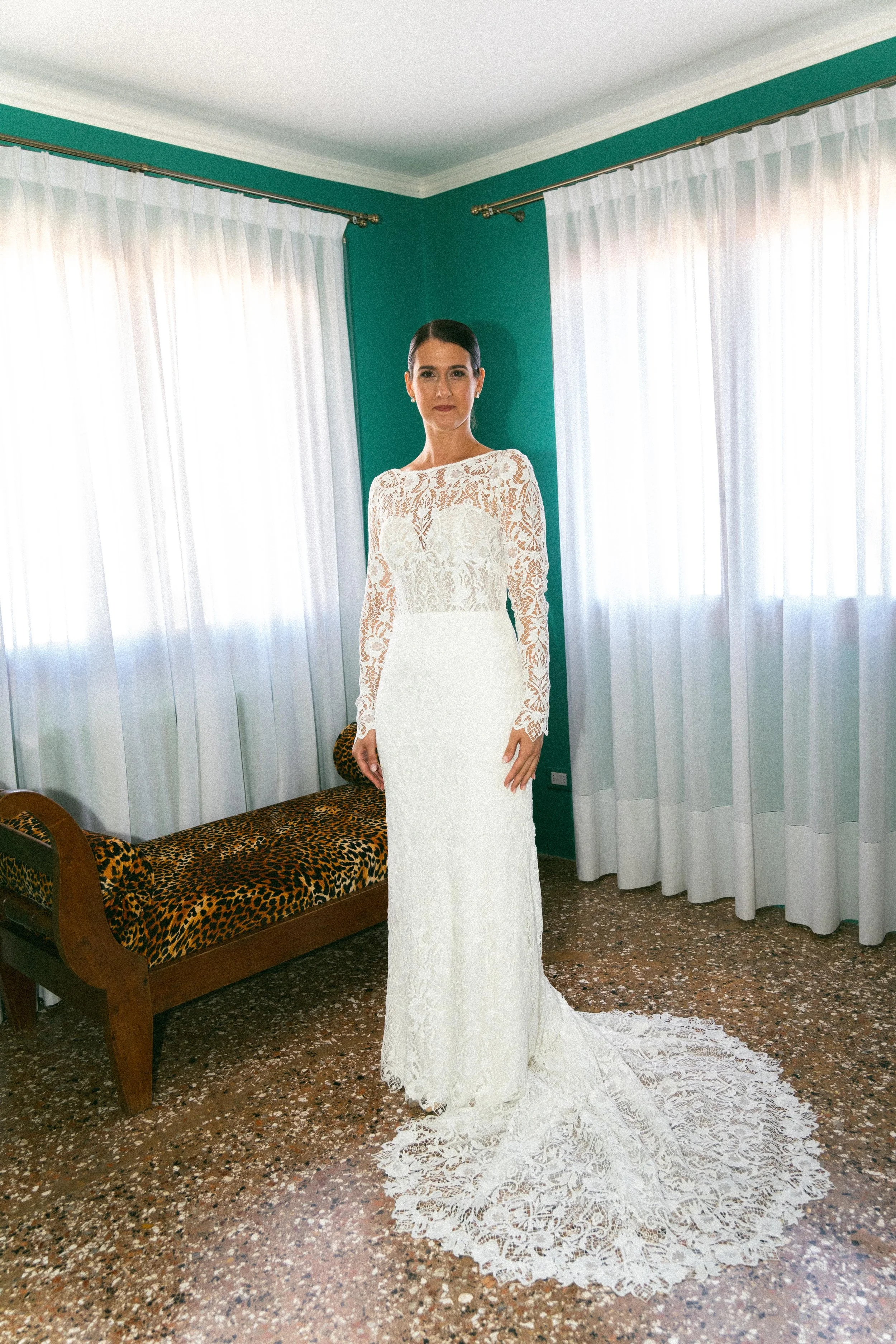 Una donna con un abito da sposa in pizzo bianco lungo e aderente, con capelli raccolti, si trova in una stanza con pareti verdi e tende bianche, davanti a un muretto con un'imbottitura con motivo leopardato.