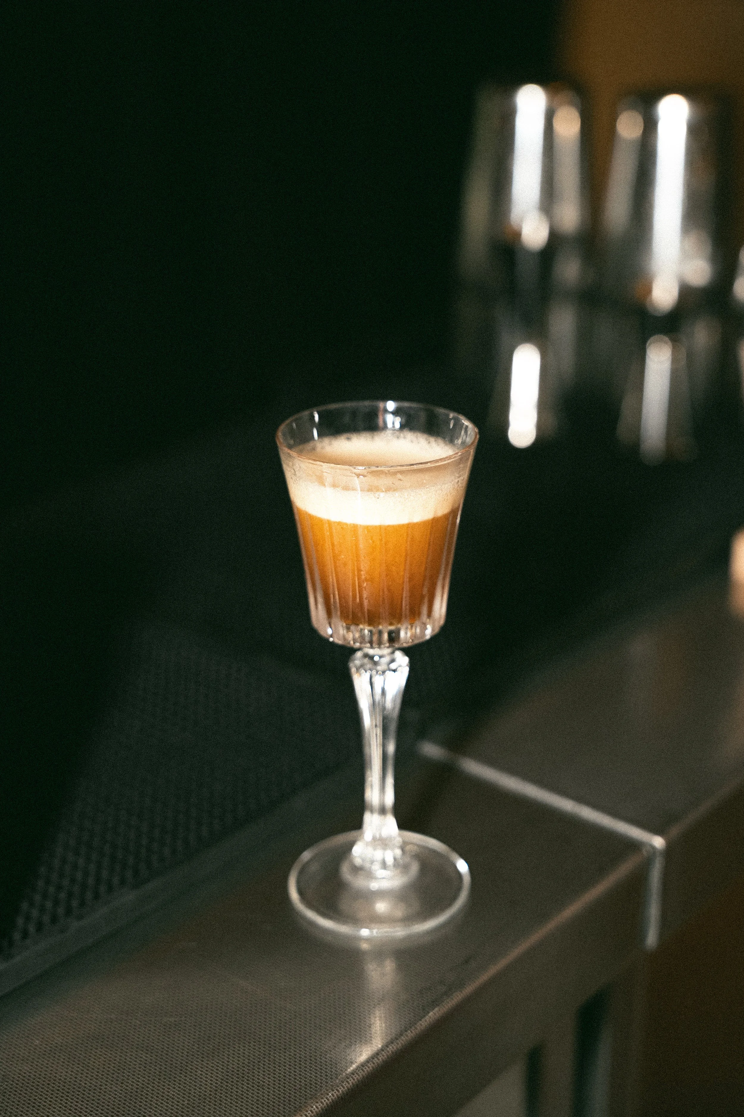 Un bicchiere da cocktail con una bevanda color marrone chiaro, decorato con una schiuma in superficie, si trova su una superficie nera in un ambiente con illuminazione calda e sfocata sullo sfondo.