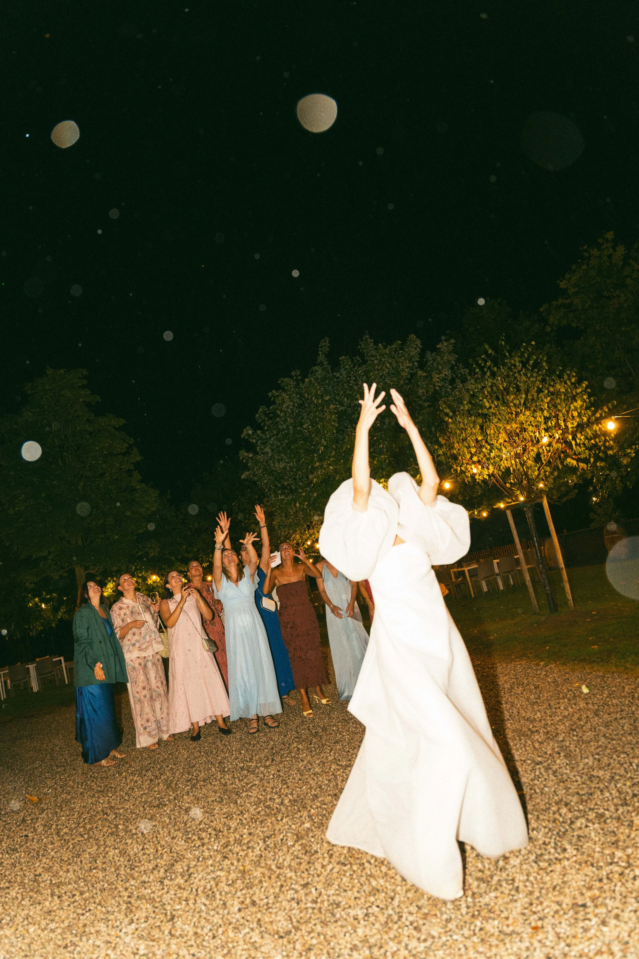 Un gruppo di persone vestite elegantemente, alcune con abiti lunghi, che partecipano a un evento all'aperto di sera, mentre qualcuna lancia o riceve qualcosa in aria, con luci calde e alberi sullo sfondo
