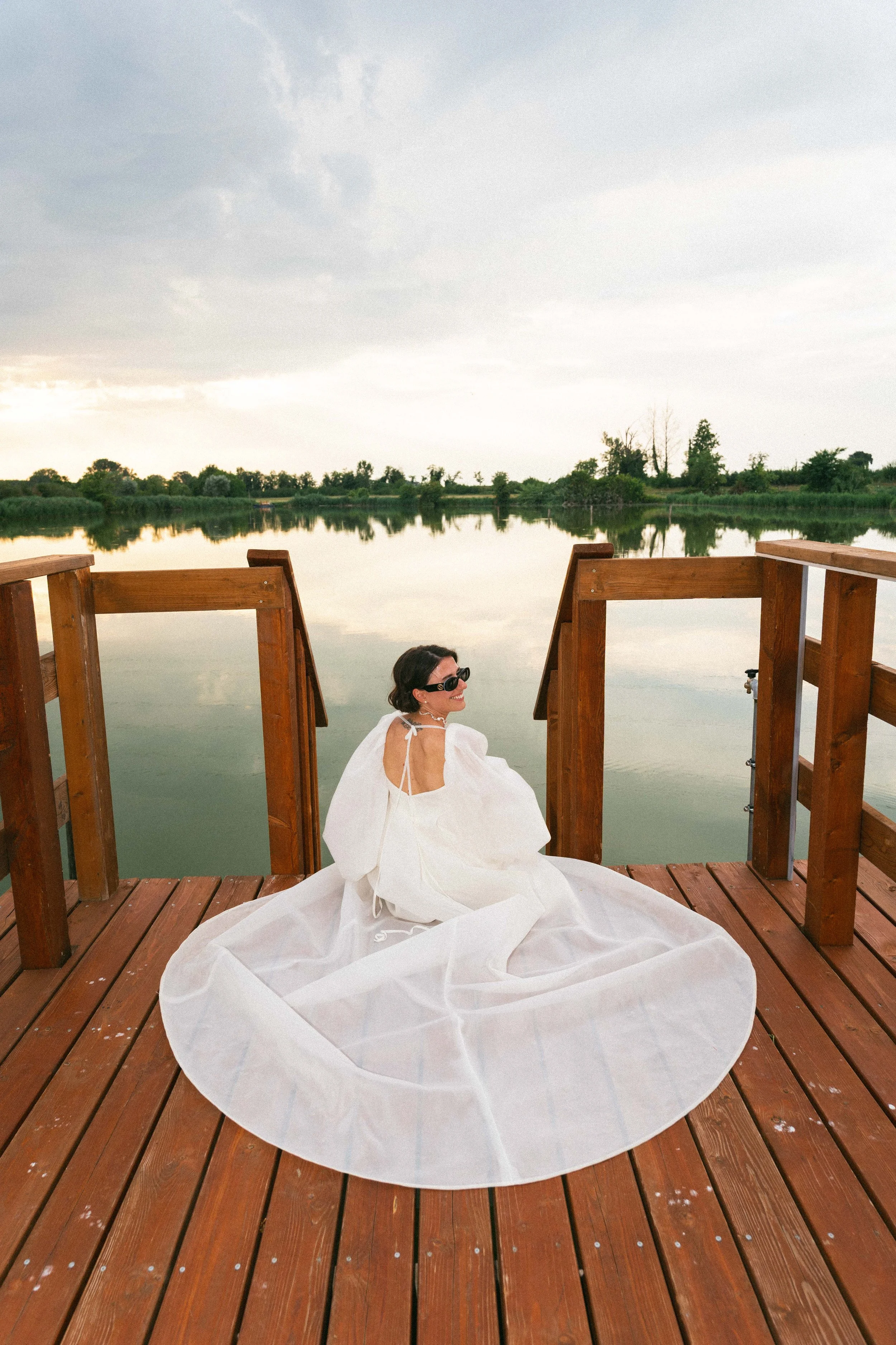Una donna sorridente con occhiali da sole indossa un abito bianco lungo e fa una posa seduta sulla ringhiera in legno di una banchina vicino a un fiume, con uno sfondo di cielo nuvoloso e natura