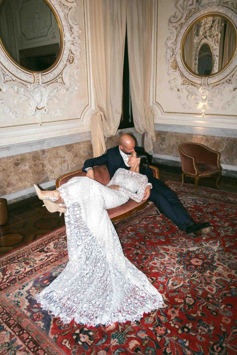 Una coppia innamorata seduta su un divano in una stanza elegante con pareti decorate, specchi rotondi e tende lunghe. La donna indossa un abito in pizzo bianco lungo e il uomo un abito scuro.