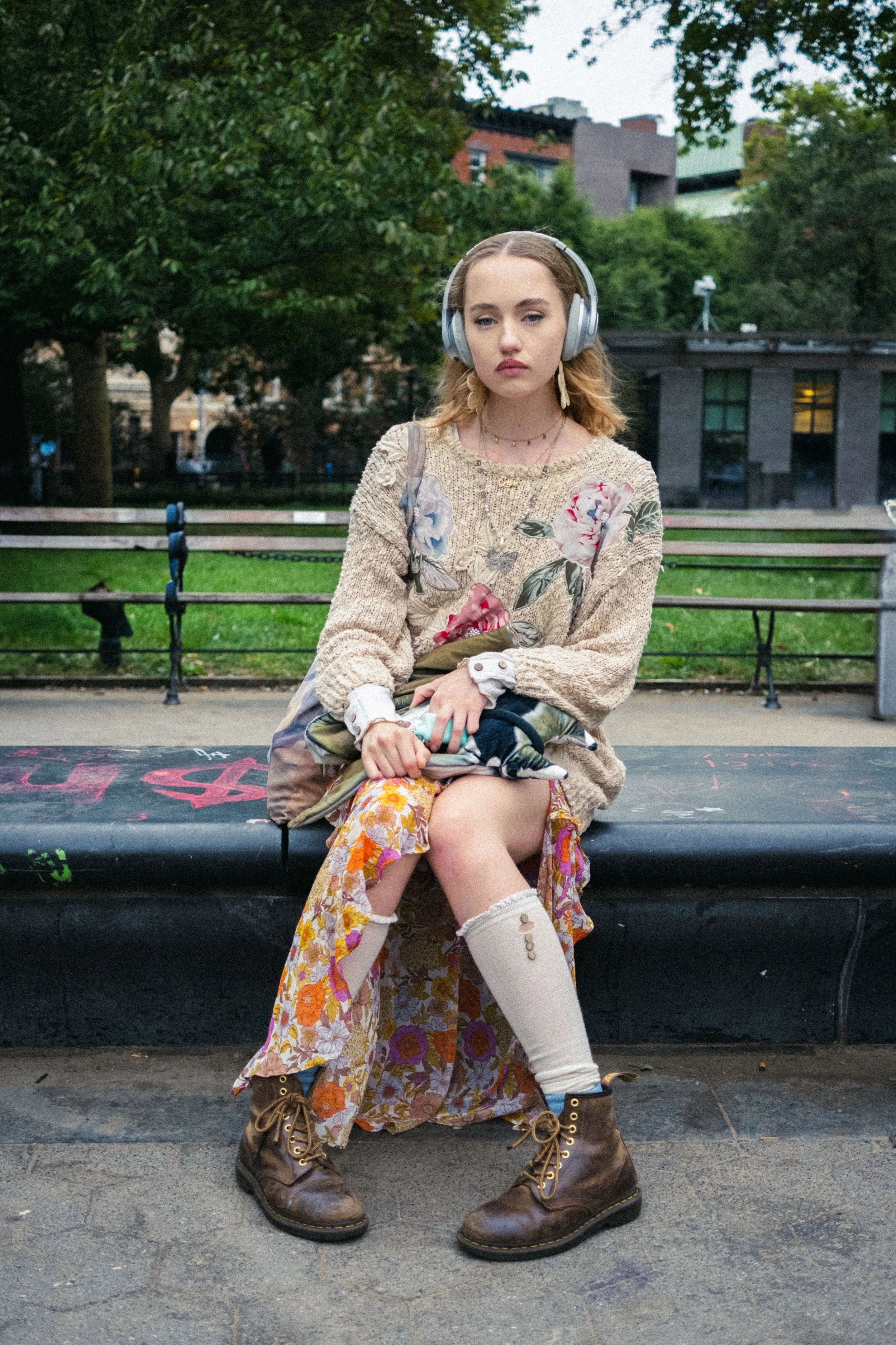 Ragazza seduta su una panchina nel parco, indossa cuffie grandi, maglione beige con motivi floreali, gonna lunga con stampa floreale, calzini acidi e stivali marroni.