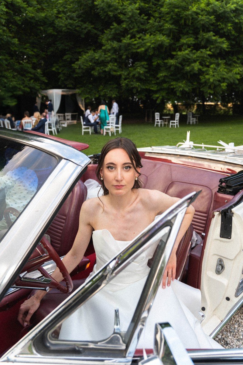 Una donna in abito da sposa seduta in un'auto d'epoca bianca con sedili rossi, in un'ambientazione all'aperto con alberi e persone sedute a tavoli in background.
