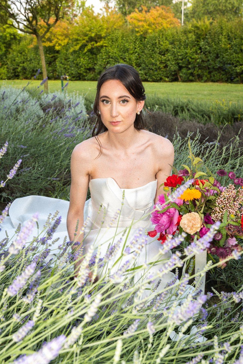 Una donna con un vestito bianco senza spalline seduta in un giardino con fiori e alberi verdi in background, con un mazzo di fiori colorati accanto.