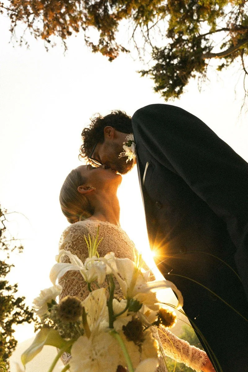 Una coppia di sposi si bacia all'aperto al tramonto, con il sole dietro, circondata da alberi e fiori.