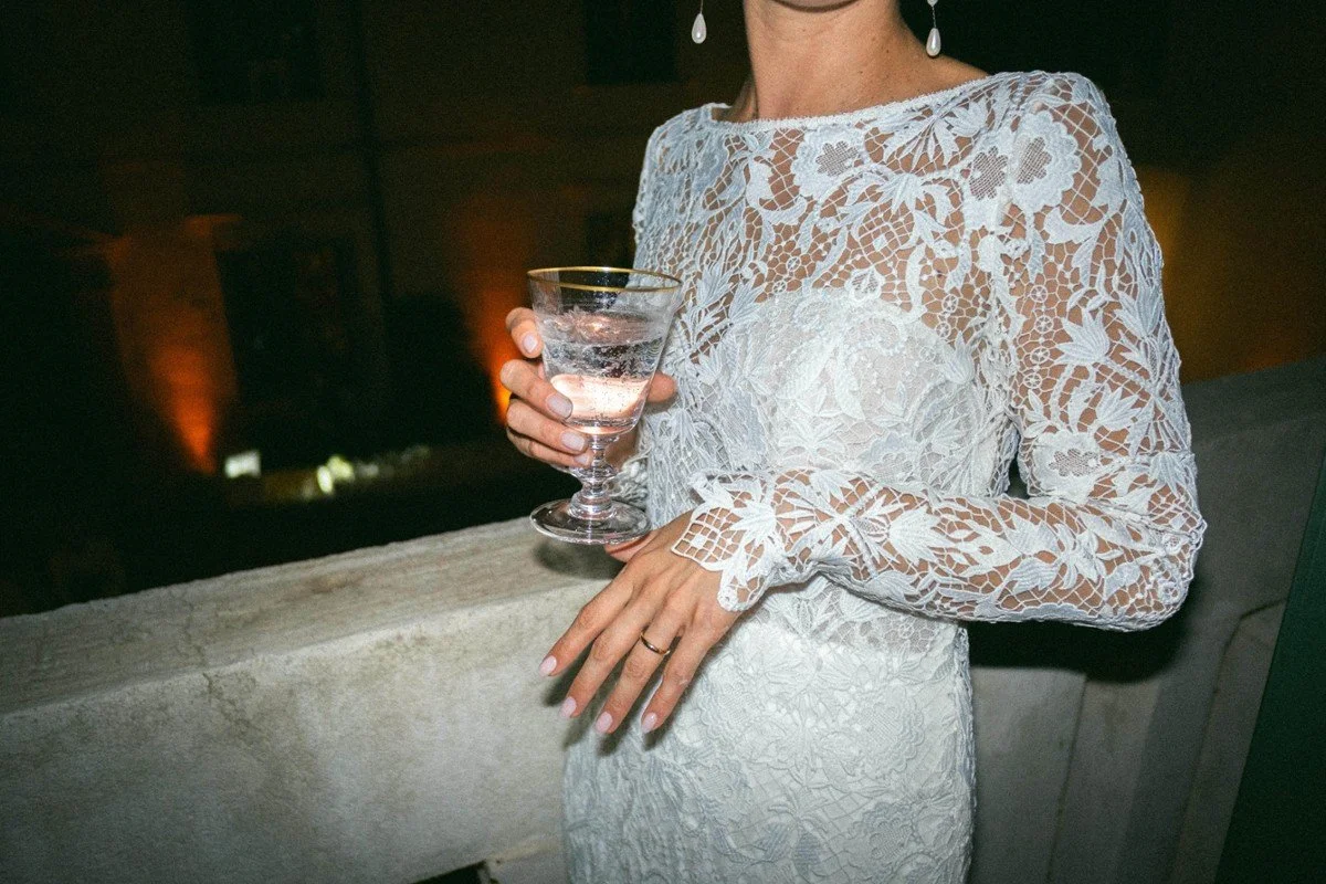 Donna con abito bianco in pizzo mentre tiene un bicchiere di drink, in un ambiente elegante.