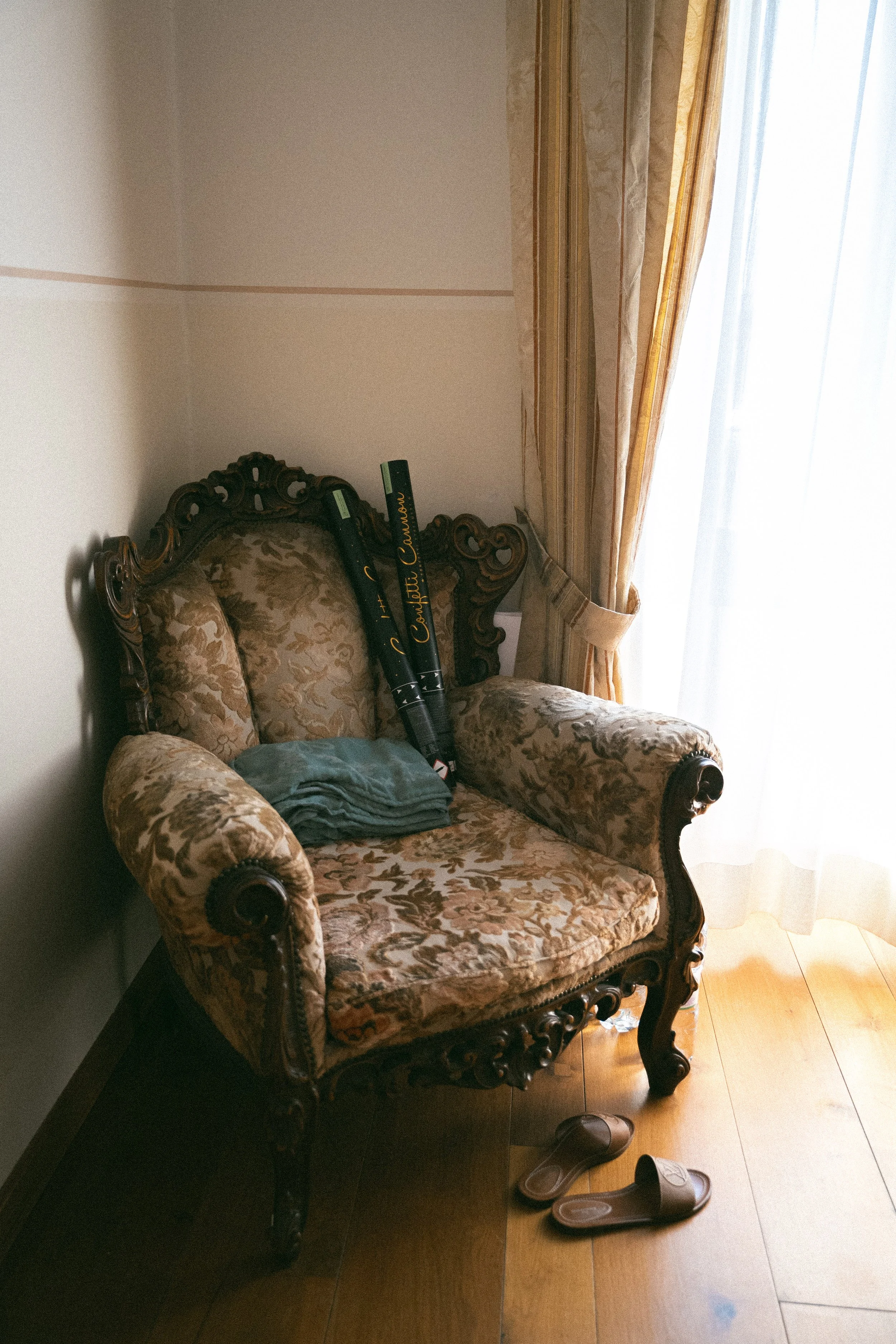 Una sedia antica in stile classico con rivestimento in tessuto damascato beige e motivo floreale, accanto a una finestra con tende dorate e cortine di pizzo, con alcuni oggetti sopra la seduta e un paio di infradito sul pavimento di legno.