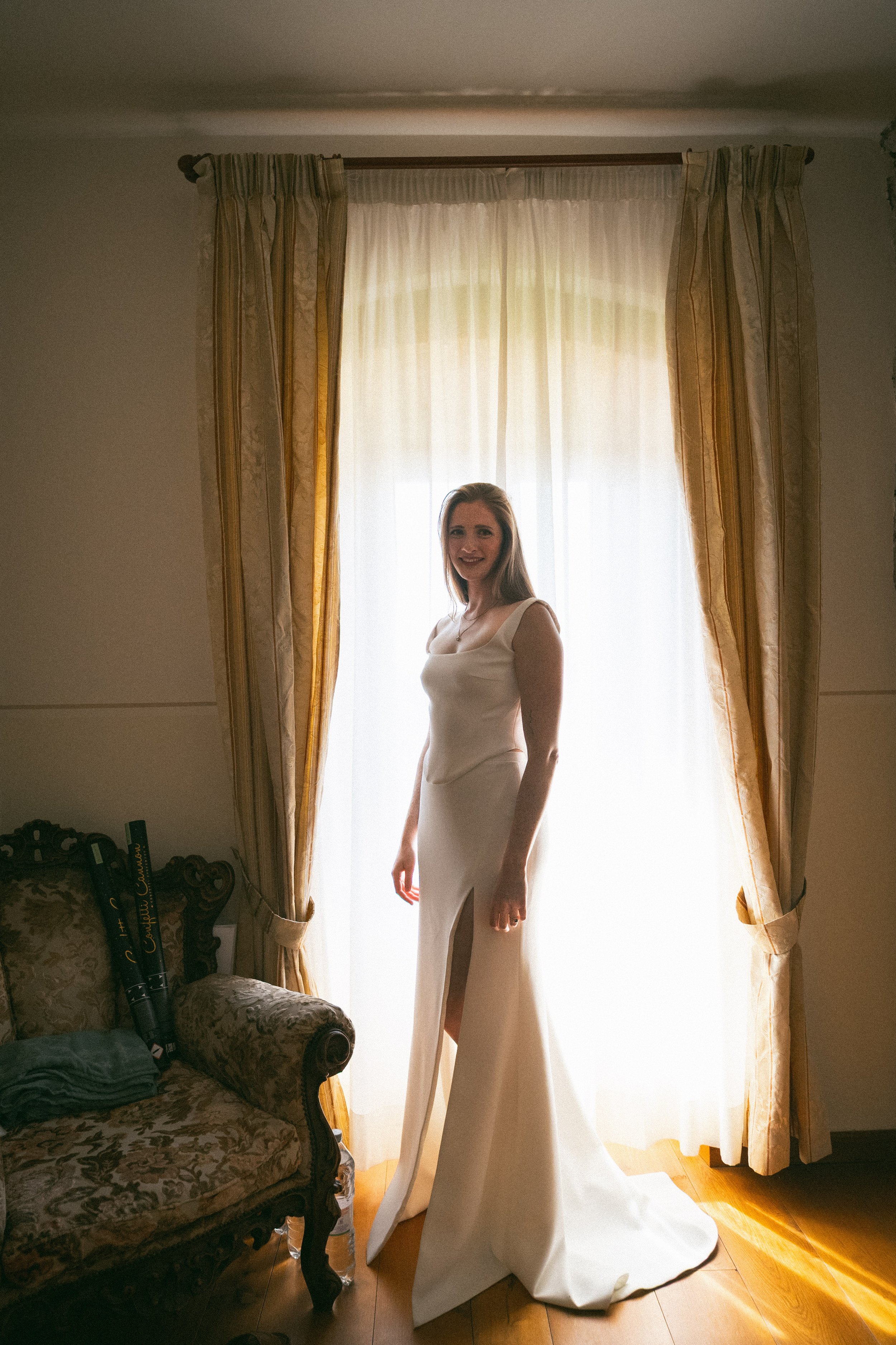 Una donna con un abito da sposa in un soggiorno davanti a una finestra con tende color avorio e oro, seduta su una poltrona in stile classico con tessuto damascato. La stanza ha pavimento in legno e luce naturale che illumina l'ambiente.